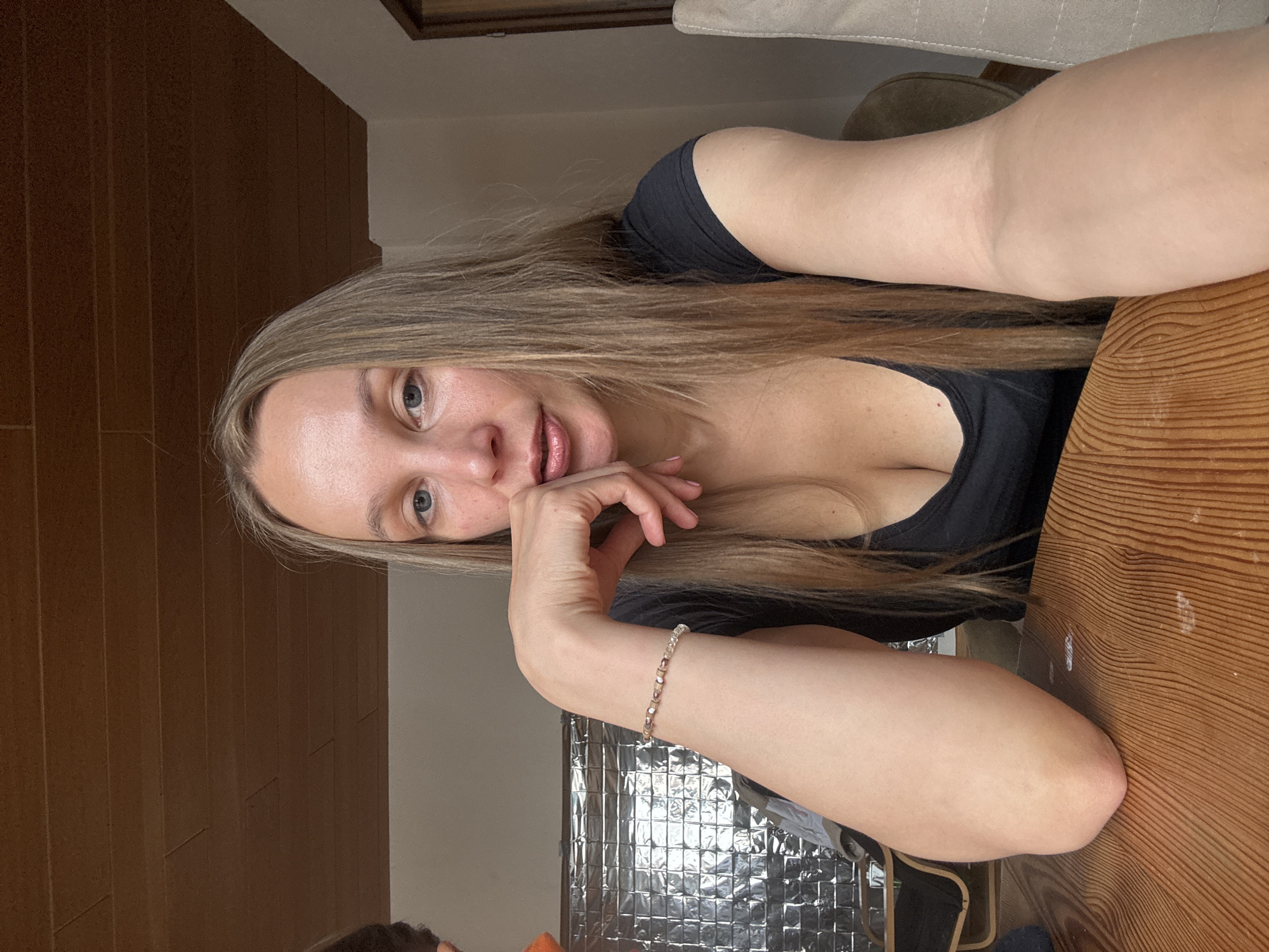 The image shows a medium shot of a woman with long, straight, light brown hair and blue eyes. She is wearing a black V-neck top and a beaded bracelet on her left wrist. Her left arm is resting on a wooden table, and her fingers are touching her chin. She is looking at the camera. The background appears to be an indoor setting with wooden paneling on the ceiling and a light-colored wall. There is a metallic-looking material on the wall behind her left arm, possibly insulation or a decorative element. To the right, a portion of a light-colored chair is visible. The lighting is natural and comes from the right side of the image. The image does not provide any specific visual cues that would allow for the determination of its location in Überherrn, Germany.
