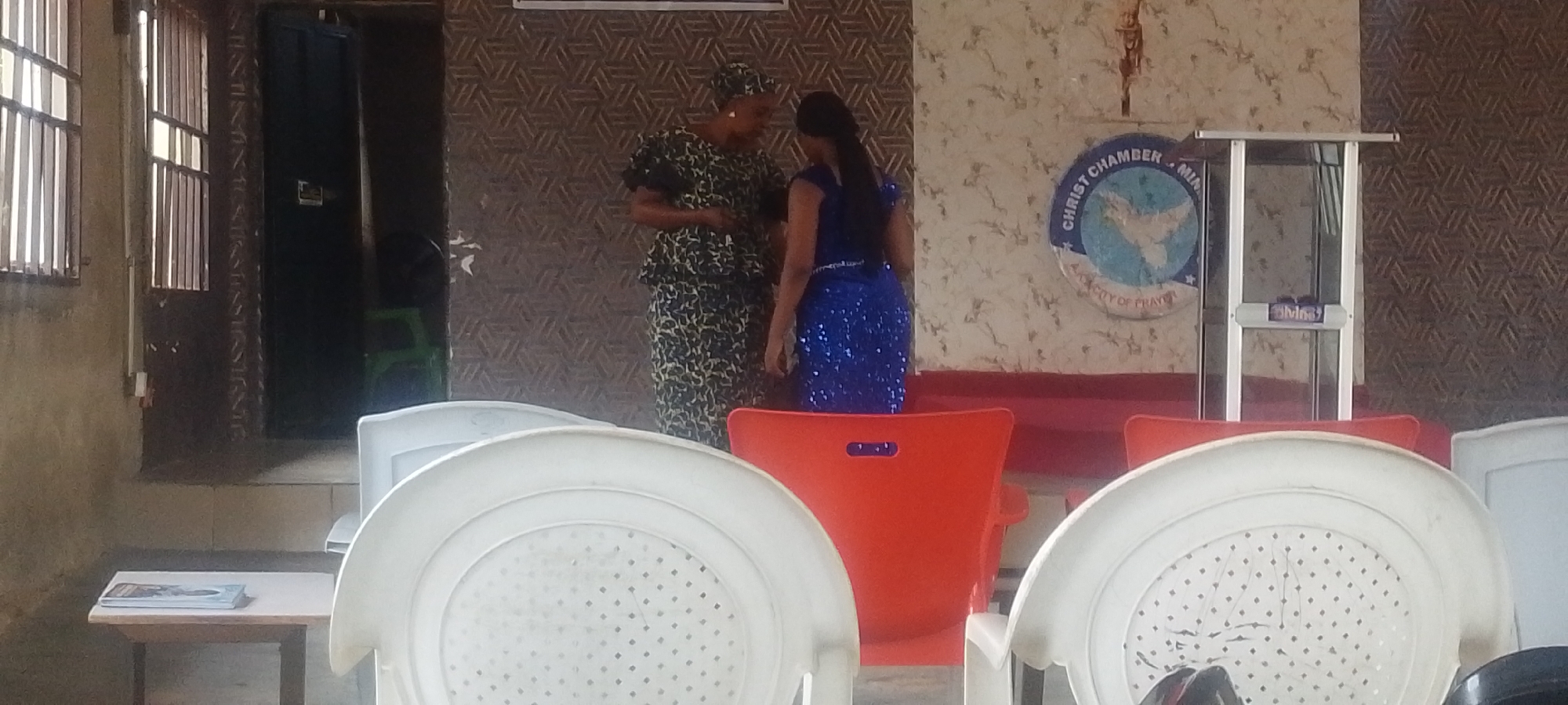 The image depicts the interior of the Christ Chamber Ministry, City of Prayer, located in Lokoja, Nigeria. Two women are standing in the foreground. The woman on the left wears a patterned dress and head covering, while the woman on the right is wearing a blue, sequined dress. Behind them is a wall adorned with a logo of the ministry, above which a small crucifix is mounted. A metal pulpit stands to the right of the logo. Several white and orange plastic chairs are arranged in rows, with their backs facing the camera. A small table with reading material is visible on the left, positioned in front of a window and a dark doorway. The walls have patterned wallpaper in the background.