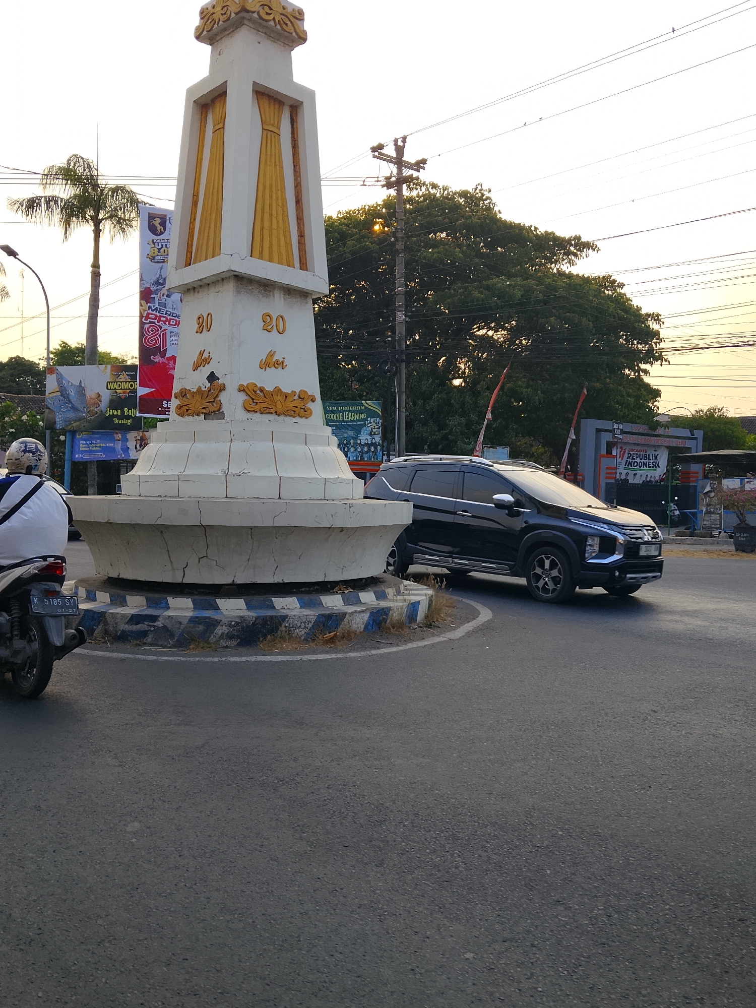 The image shows a street scene in Balun, Indonesia, dominated by a large monument and a busy intersection. The monument, in the foreground and to the left, is white with gold decorative elements and inscribed with "20 Mei" twice. A motorcycle is parked to the left of the monument, with a person wearing a white shirt and a helmet sitting on it. The road is paved, and there are various advertisements and signs in the background, including one that reads "REPUBLIK INDONESIA." A black SUV is driving through the intersection, and a tall tree is visible behind the monument. Power lines crisscross the sky, and the lighting suggests it is late afternoon or early evening. The environment appears to be an urban or semi-urban area with traffic and pedestrian activity.