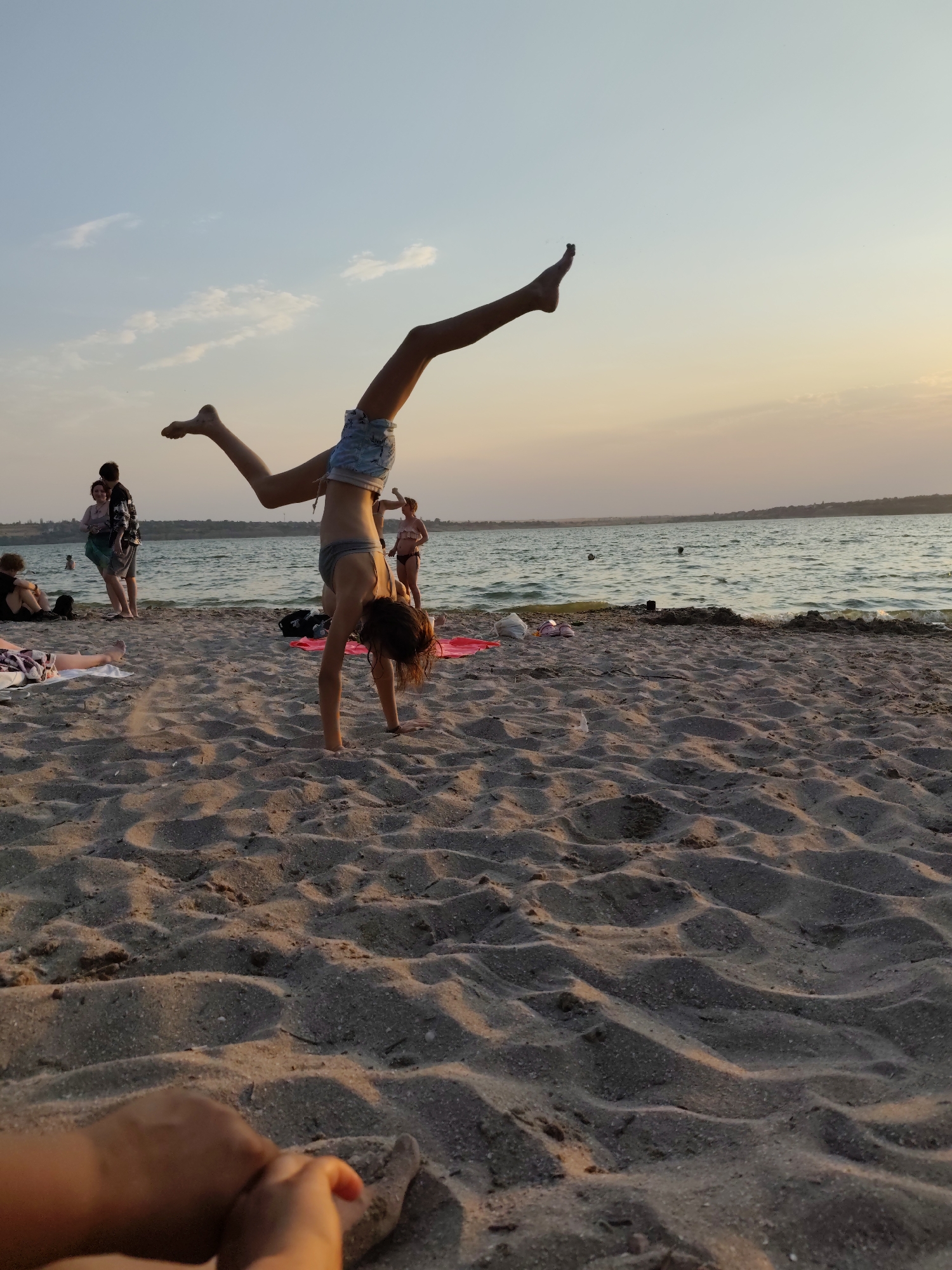 The image shows a beach scene with a young woman performing a handstand in the foreground. She is wearing a grey bikini top and patterned shorts. Her legs are extended upwards, with one leg bent. The beach is sandy, with footprints and sand ripples visible. In the background, a body of water, likely a river or the sea, stretches to the horizon. Several people are visible near the water, some standing and others sitting on the sand. The sky is a soft gradient of blue and orange, suggesting either sunrise or sunset. The overall atmosphere is relaxed and recreational. The location is described as the city of Mykolaiv, Ukraine, a port city on the Southern Bug River, which flows into the Black Sea.