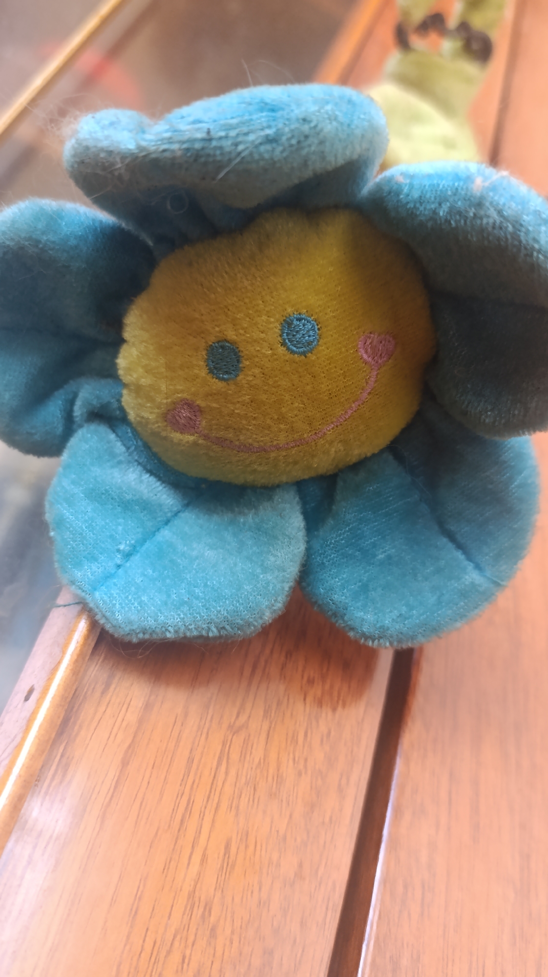 A close-up shot shows a blue plush flower toy with a yellow center. The flower has large, soft blue petals and a yellow face embroidered with two blue dots for eyes, pink blush marks, and a pink smiley mouth. The toy is resting on a wooden surface, possibly a table, with a blurred background that suggests an indoor setting. The lighting is warm and diffuse, indicating it might be daytime or a well-lit room. The focus is on the toy, highlighting its fuzzy texture and cheerful expression. There is no visible text in the image. The overall mood is playful and innocent.