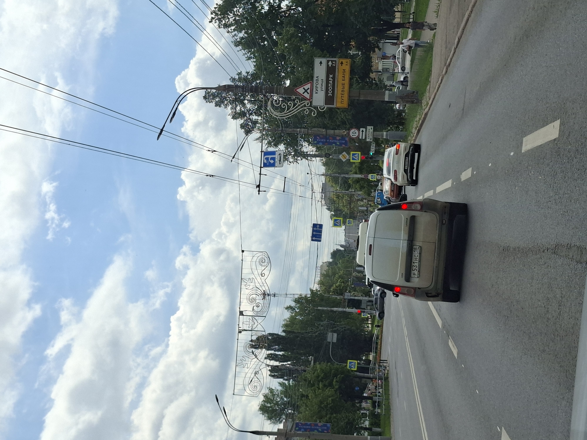 The image captures a street scene in Izhevsk, Russia, likely on a cloudy day with patches of blue sky. A white van is in the foreground, traveling down a multi-lane road. Behind it, a white Mercedes sedan is also moving in the same direction. The road is asphalt, with clear lane markings.
Along the right side of the road, there is a sidewalk with pedestrians. Buildings and trees line the street. Overhead, electrical wires crisscross the sky, supported by poles. Several traffic signs are visible, including directional signs indicating "KIROVA street", "ZOO PARK", and "PUTEVYE BANI", as well as traffic signal lights and pedestrian crossing signs.
The van has a license plate that reads "P 531 HC 18". Another vehicle's license plate is partially visible on the Mercedes, showing "B 102 TE 777". The overall atmosphere is that of a typical urban environment during daylight hours.