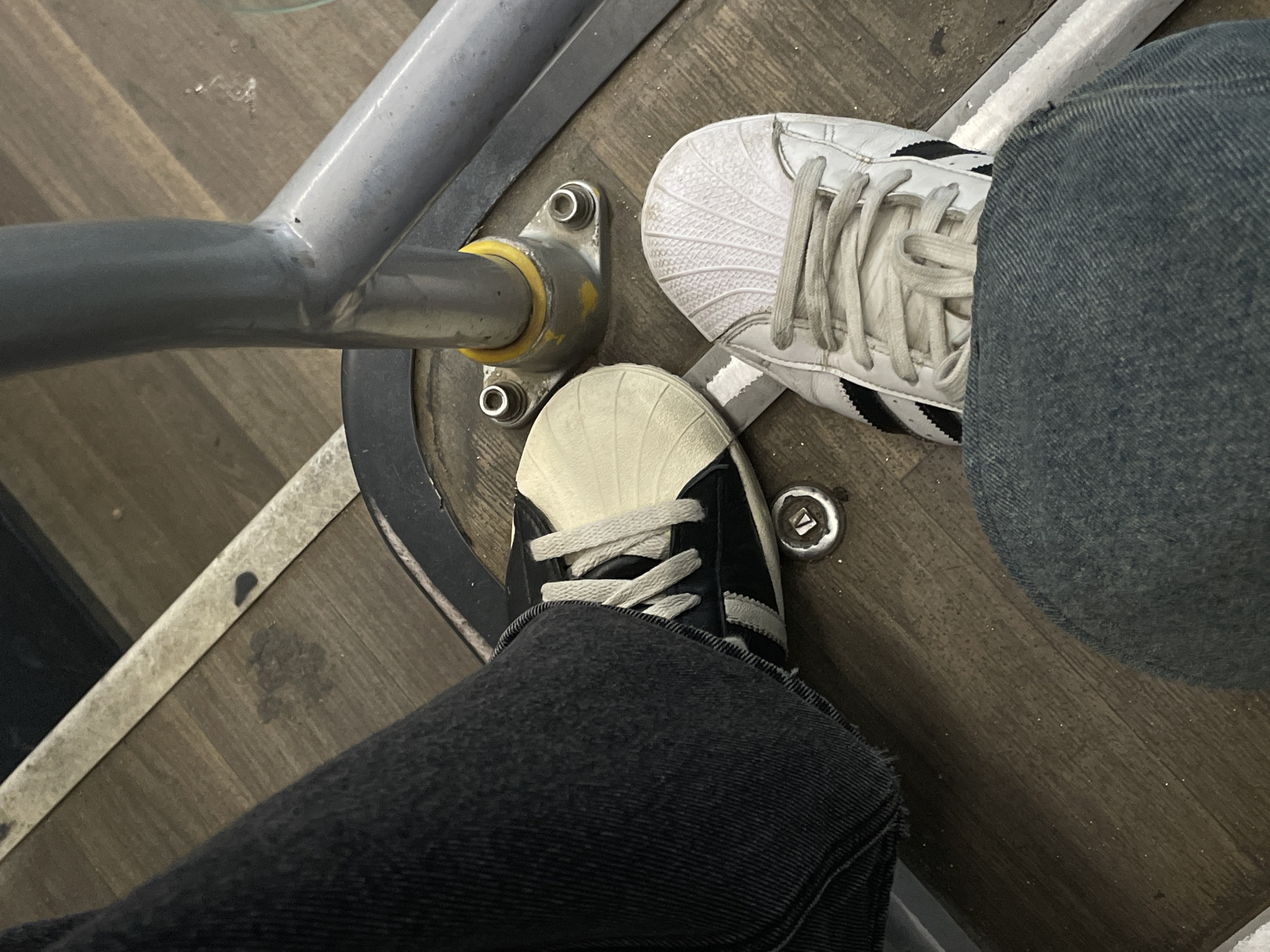 The image shows a close-up, overhead view of two pairs of feet, wearing Adidas Superstar sneakers, on a wooden floor. One person is wearing white Adidas Superstars with black stripes, and dark blue denim jeans. The other person is wearing black and white Adidas Superstars with white laces, and dark grey denim jeans. Both individuals appear to be seated or resting, as their feet are positioned on the floor, with one leg crossing over the other. A metal railing, possibly from a bus or train, runs diagonally across the top left of the frame. The floor is made of wooden planks, and there are several circular metal fixtures visible on the floor, likely part of the seating or structure of the vehicle. The overall lighting suggests an indoor setting, possibly during the day. There is no visible text in the image. The context suggests this scene is taking place in Yakovlevskoye, Russia.