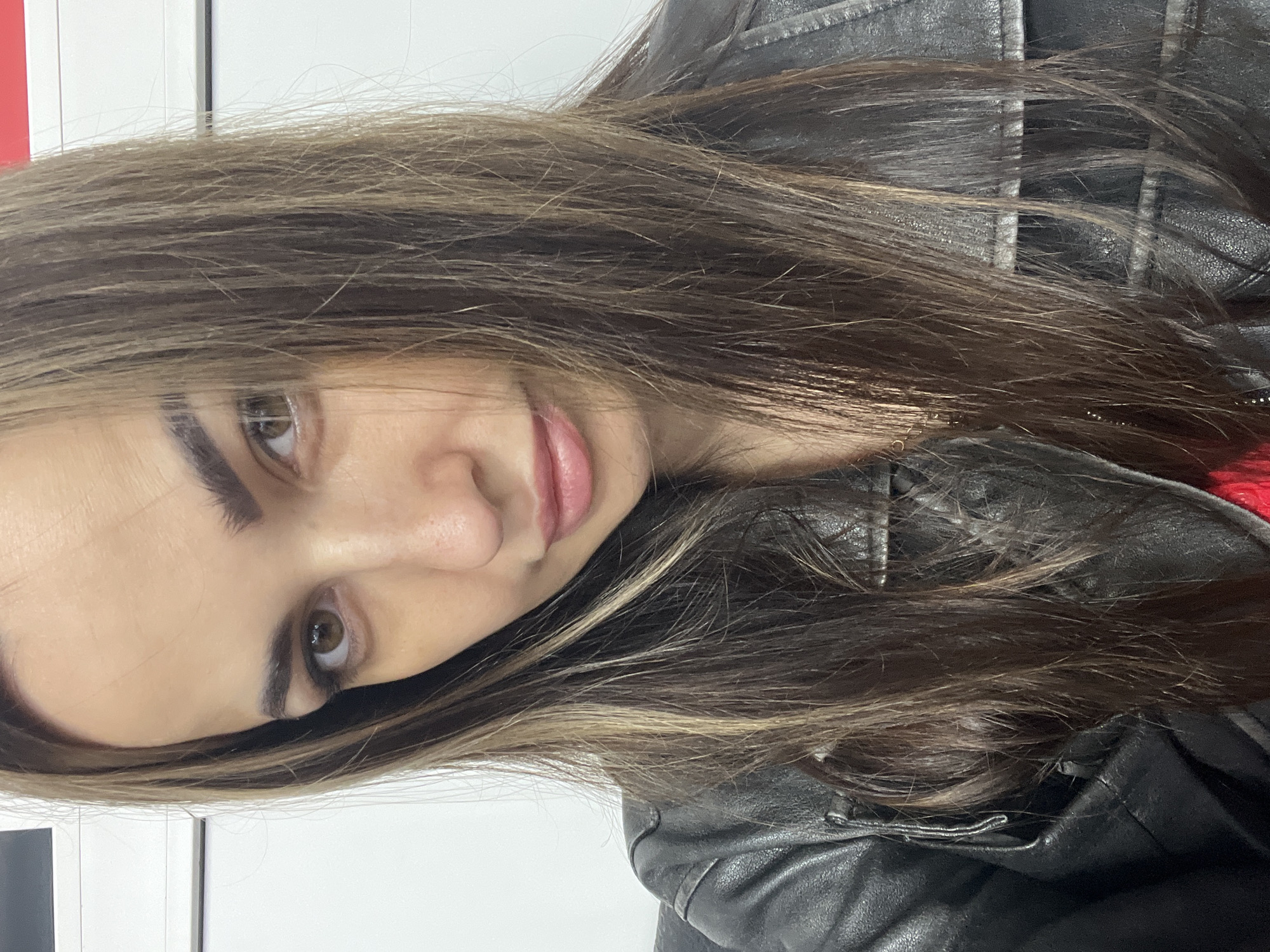 A close-up, chest-up portrait shows an adult female gazing directly at the camera in an indoor setting in Unknown, Unknown. She has long, straight to slightly wavy dark brown hair with lighter, possibly caramel-toned, highlights that falls over her shoulders and partially across her face. Her eyes are brown, and her eyebrows are dark and well-defined. Her lips are full and lightly colored, slightly parted in a subtle, faint smile. She wears a black leather-style jacket, with a glimpse of red fabric visible underneath on her right side. The background consists of plain white walls, with a distinct horizontal red strip and a dark line visible on the upper right side.