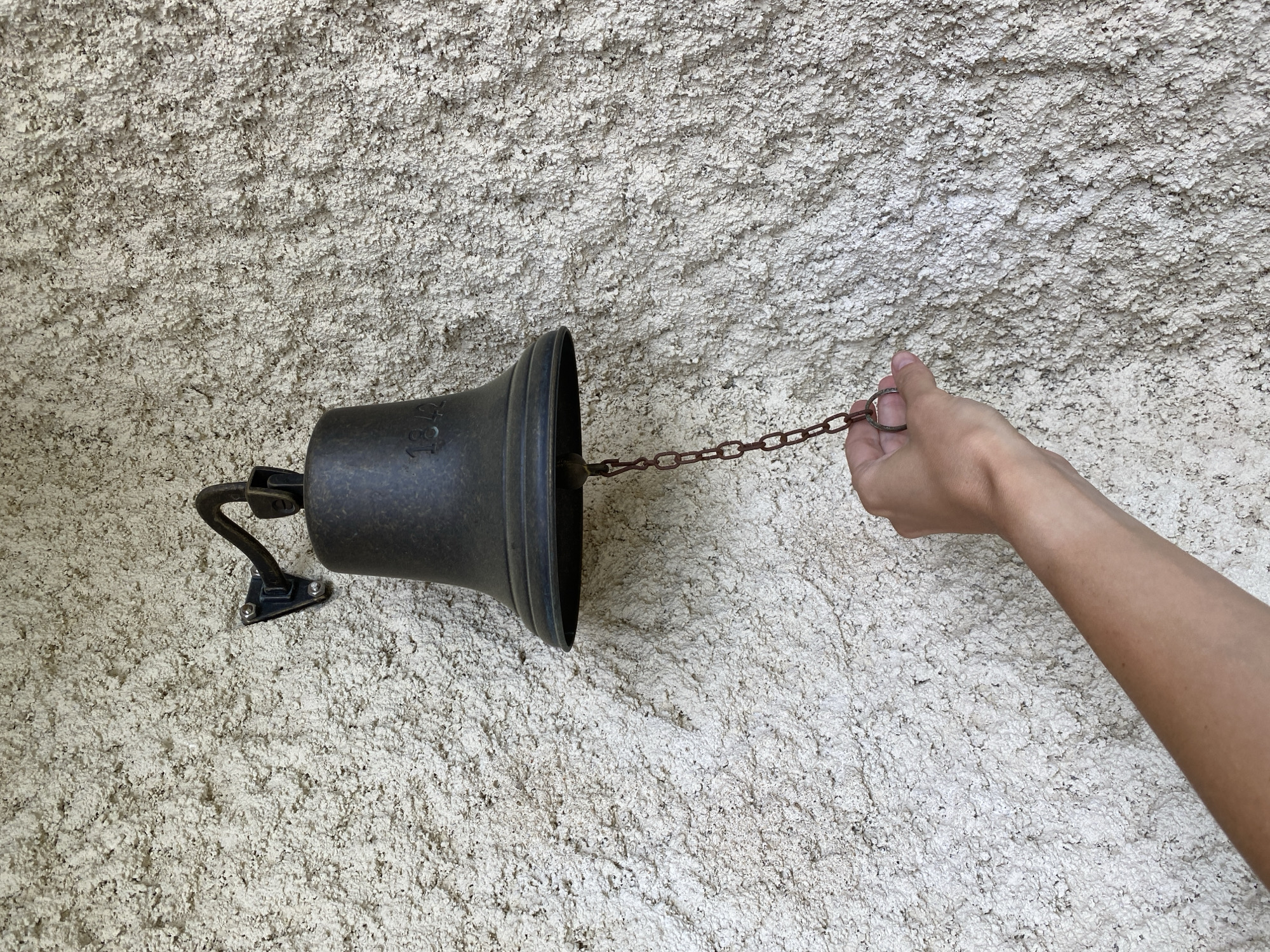 A close-up, eye-level shot captures a human hand pulling a chain to ring a bell mounted on a textured wall. The scene is illuminated by natural light, suggesting daytime.
The primary subjects are a person's light-skinned hand and forearm, and a dark-colored metal bell. The hand is gripping the last link of a rustic, dark metal chain, extending from the bell towards the right side of the frame. The bell itself is a classic, flaring shape, cast in a dark, possibly antique bronze or iron material. It is securely attached to the wall via a black metal bracket with two visible screws. Faint numbers "184" are visible on the side of the bell, with a partially obscured fourth digit.
The background is a highly textured wall, appearing to be made of coarse stucco or rough-cast plaster. Its color is a light, off-white or beige, with shadows and variations indicating its uneven, granular surface. The image emphasizes the tactile interaction of ringing the bell against this rustic backdrop. The location context is Courpière, France.