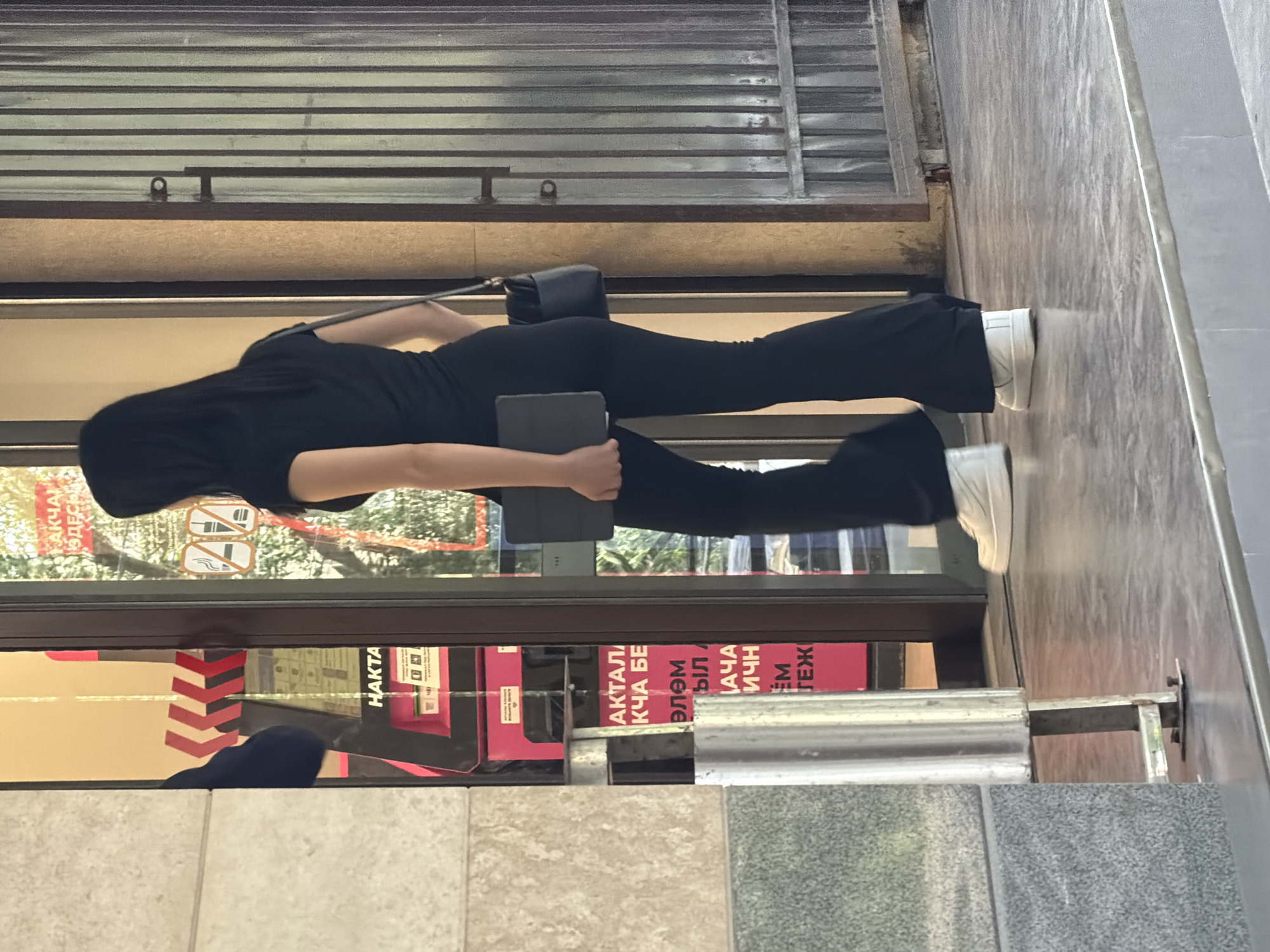 A woman in black clothing and white sneakers is walking past a storefront. She is carrying a dark tablet and a black purse. The storefront has a glass door and a metallic grate. There is a pink ATM or vending machine visible inside the storefront. The ground is paved with gray tiles.