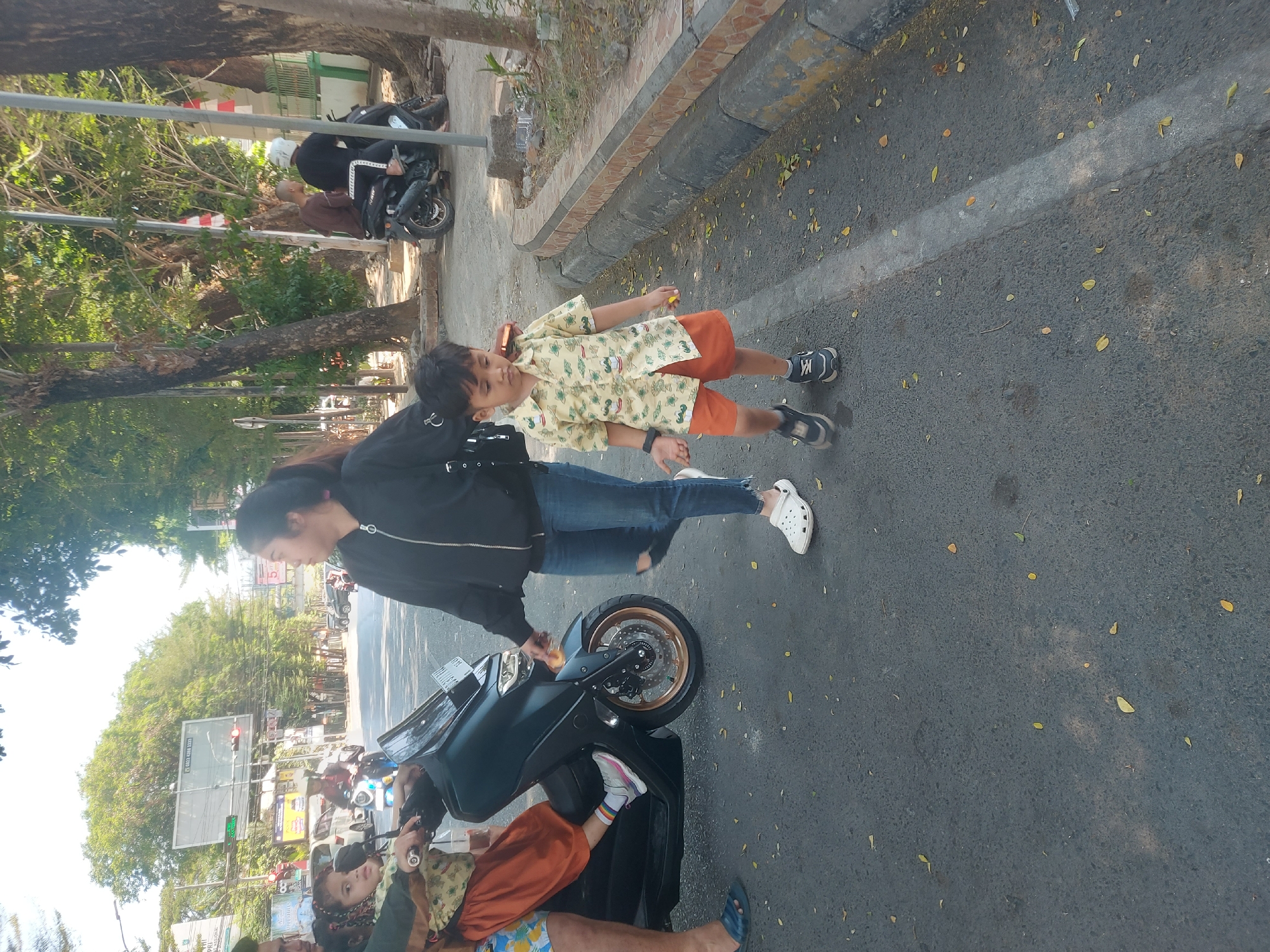 In Makassar, Indonesia, a group of individuals are gathered near a dark-colored scooter on a sunlit, paved street. A young boy, approximately five years old, walks towards the right side of the frame, wearing a short-sleeved, patterned yellow shirt and orange shorts, with dark sneakers on his feet. To his left, a woman in a black jacket and ripped blue jeans walks in the same direction, looking to the side. Seated on the scooter is a young girl, wearing an orange skirt and a light-colored, patterned top, her hair in pigtails, and her feet resting on the scooter's footrest. Another person's legs are visible behind the girl, wearing blue sandals. In the background, traffic lights, trees, and buildings suggest an urban setting. Further in the background, two men on a motorcycle are visible. The ground is asphalt, scattered with fallen leaves, and a concrete curb runs along the right side of the street.