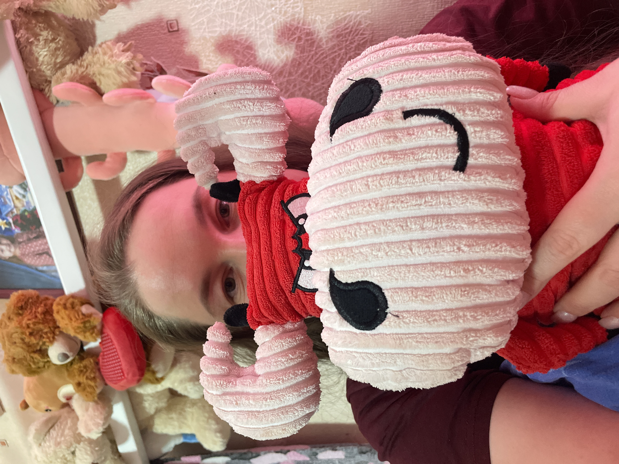 A person is lying down, holding a plush toy in front of their face. The toy has a white, cylindrical body with a smiling face stitched on it in black. It also has red arms and small white horns. The person appears to be indoors, surrounded by other plush toys. There is no information in the image to indicate that the location is the city of Izhevsk, Russia.
