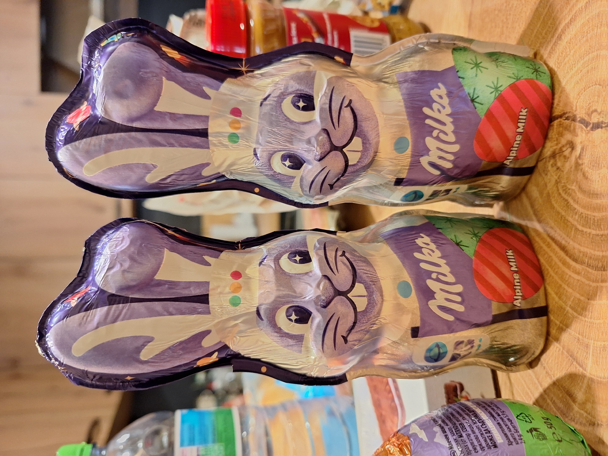 Two wrapped Milka chocolate bunnies are positioned on a wooden surface. The wrappers feature a cartoon rabbit face and the brand name "Milka" along with "Alpine Milk" text. The bunnies are in focus, while the background is blurred, revealing items such as bottles and food packaging. There are no people visible. The setting is a domestic environment. There is no indication of Varna, Bulgaria.