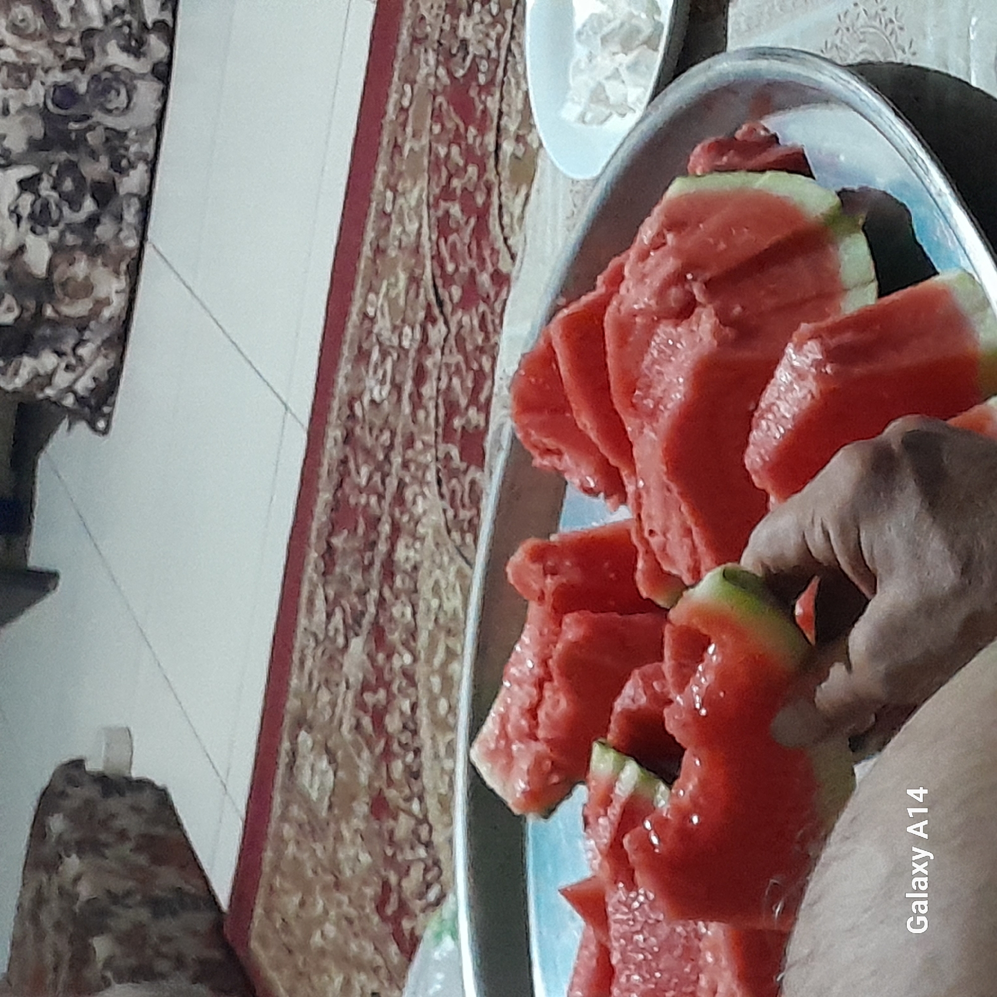 A close-up shot shows sliced watermelons on a metal platter, with a hand reaching to pick up a slice. In the background, a patterned rug is visible on the floor. A small white bowl containing what appears to be sugar cubes or candy is also present. The "Galaxy A14" watermark is visible in the bottom left corner. The image is likely from a domestic setting in Khan Yunis, Palestinian Territories.