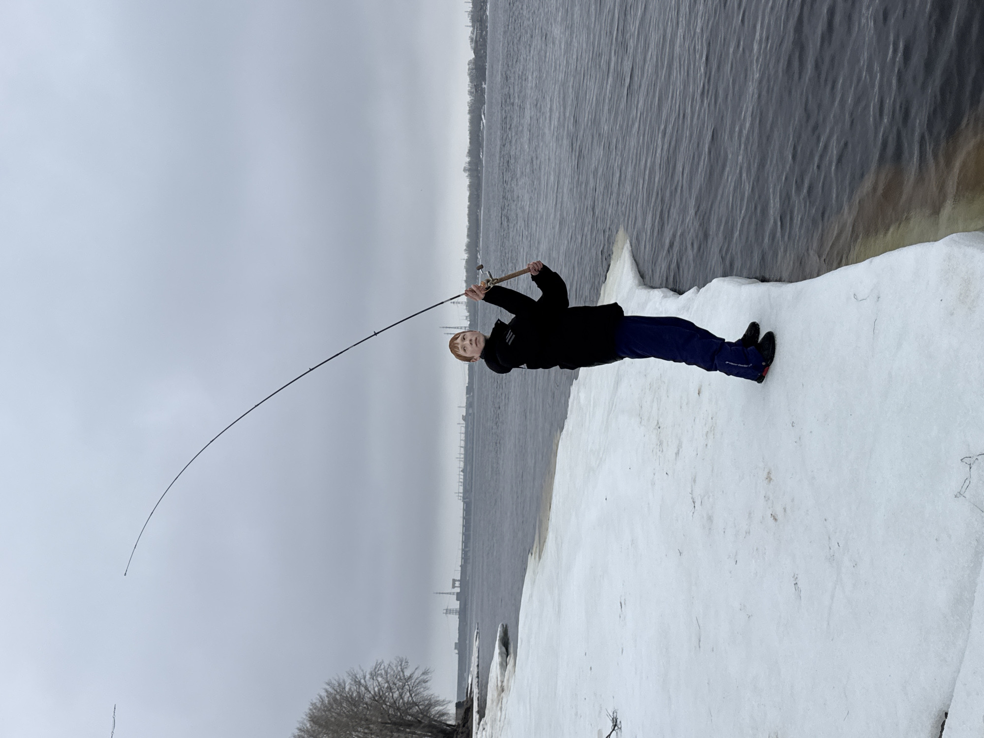 A young person is fishing on a snowy bank in Chaikovsky, Russia. The scene is overcast. The individual is wearing a dark jacket and blue pants with dark shoes. They are holding a fishing rod that is bent, suggesting there may be a fish on the line. The backdrop includes a body of water and distant industrial buildings on the opposite shore. Patches of snow and ice are present on the ground.