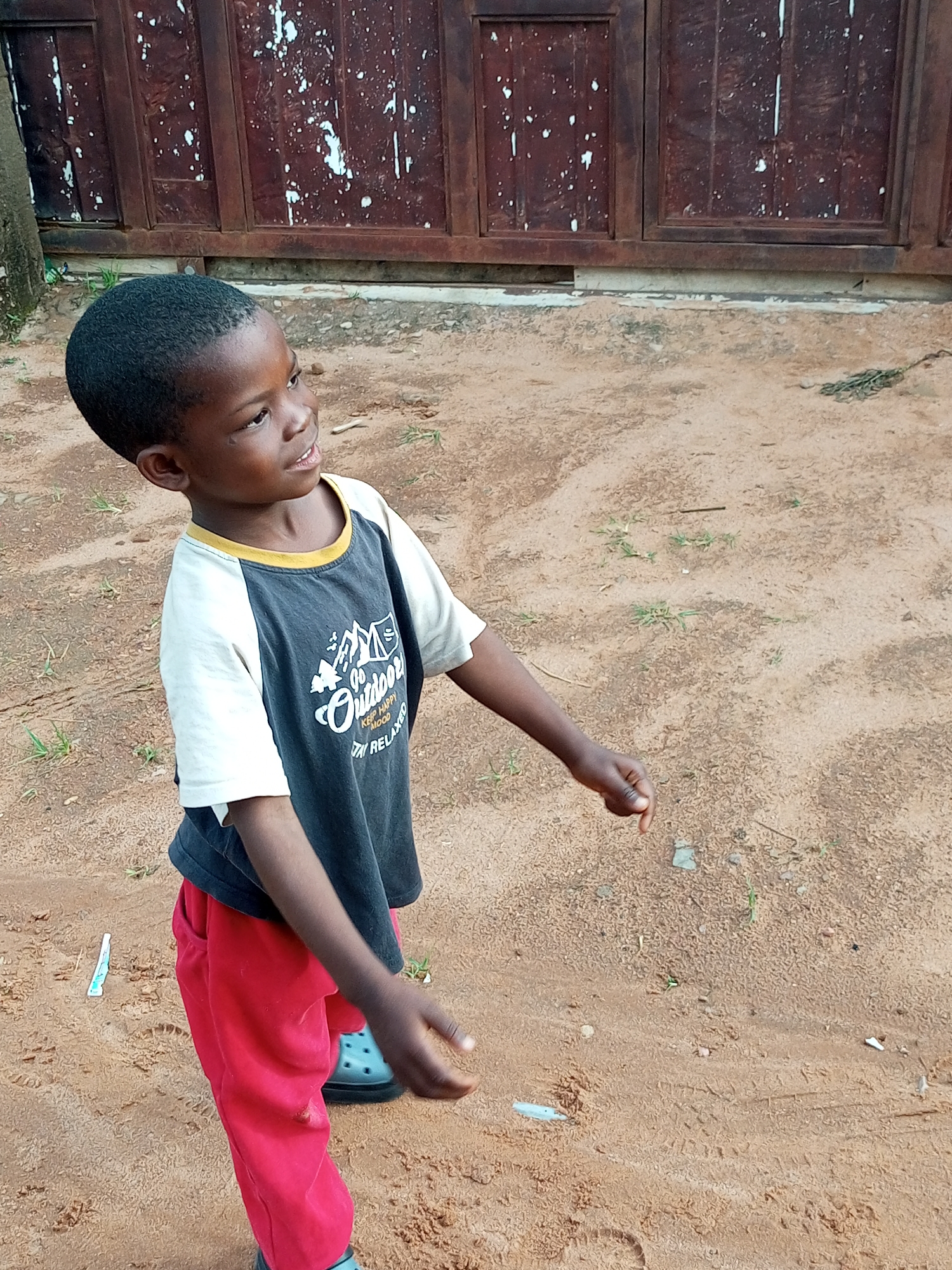 In Tempure, Nigeria, a young boy with dark skin and short, dark hair stands on a sandy surface. He is wearing a dark t-shirt with white sleeves and a yellow collar, and bright red pants. The t-shirt has a graphic design that includes the word "Outdoor" and images of mountains. His left hand is extended forward and slightly downward, while his right arm is bent at the elbow and his hand is also extended. The boy is looking upwards and to his right. Behind him is a wooden fence or door with a dark, weathered appearance, splattered with white marks. The ground around him is composed of sandy soil with scattered blades of grass and small stones. A blue shoe is visible on his left foot. A small, white object resembling a cigarette butt or wrapper lies on the sand to his left. The overall scene suggests an outdoor setting in a residential or common area.