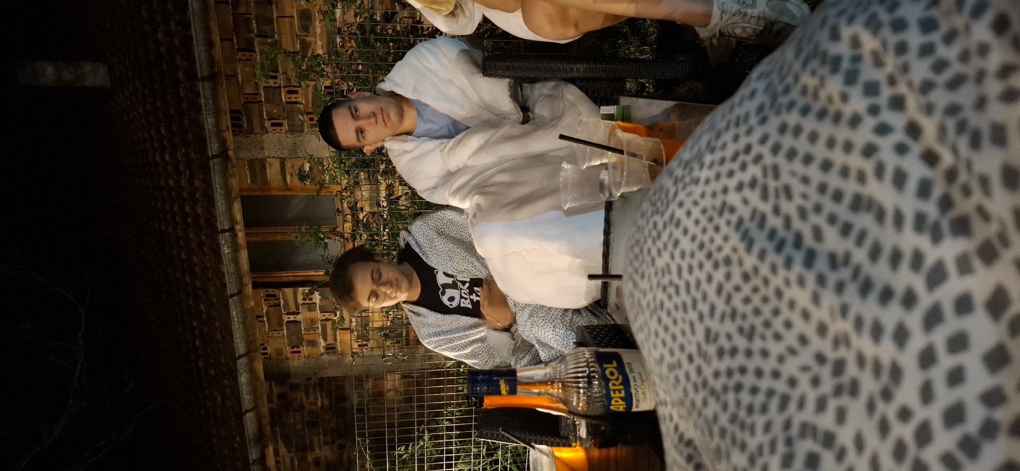 In Lobosh, Bulgaria, two young men are seated outdoors at night. The man on the left is wearing glasses and a black t-shirt with a panda logo, and is wrapped in a grey and white patterned blanket. The man on the right, wearing a light blue shirt, is covered in a white blanket. They are seated on chairs at a glass-top table. On the table are several plastic cups with straws, some containing drinks, and a bottle of Aperol. Behind them is a brick building with a tiled roof and a metal fence. The setting appears to be a backyard or patio area.