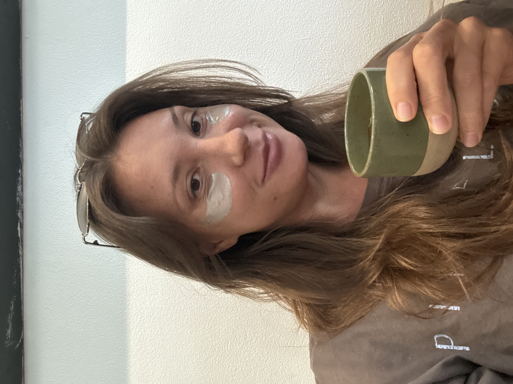 A woman with long, light brown hair poses for a selfie while holding a ceramic mug with green and beige colors. She has round sunglasses perched on her head and two round, white eye patches on her face, one on her forehead and the other on her cheek. The background includes a white wall and a dark, indistinct object on the left. The woman appears to be wearing a brown t-shirt with a white graphic. The setting is identified as Kuta Selatan, Indonesia.