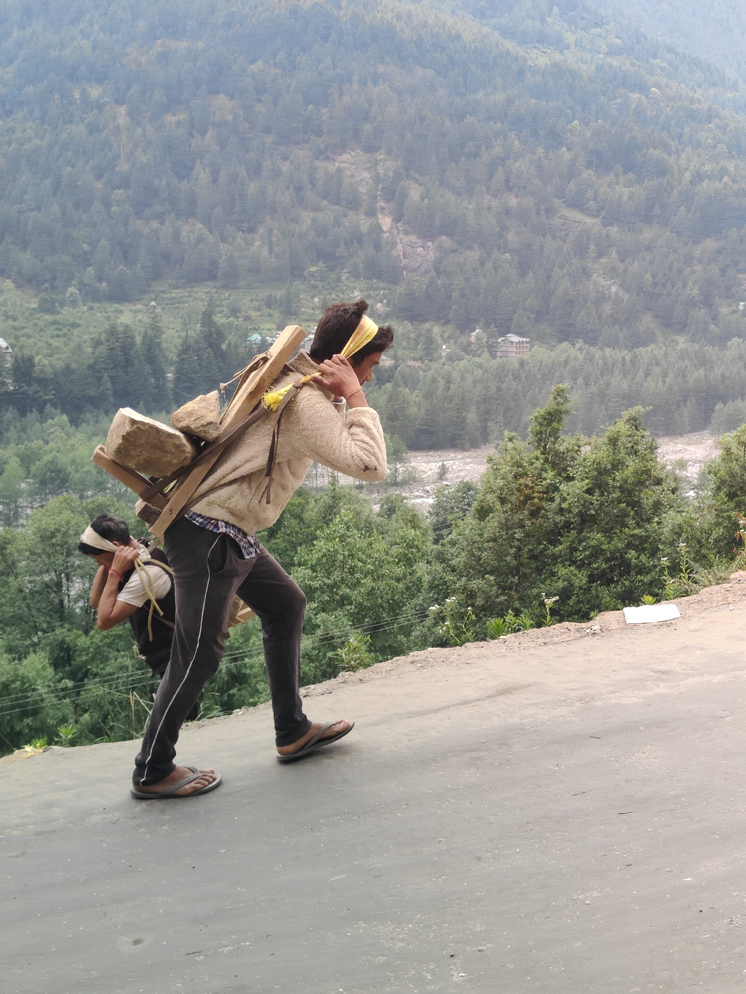 The image depicts two individuals carrying heavy loads on their backs, walking up a paved road that ascends a hillside. The man in the foreground, wearing a beige sweater and dark pants, is carrying a large wooden frame loaded with stones. He has a yellow bandana tied around his head. Behind him, a second individual, partially visible, is also carrying a load and wearing a white shirt and a head covering. The background is dominated by a dense, forested mountain slope, with a river visible in the distance and some small structures nestled within the trees. The environment appears to be a mountainous region, likely in or near Vashisht, India, characterized by its natural landscape and rural or semi-rural setting. The lighting suggests it is daytime, possibly overcast or during a hazy period.
