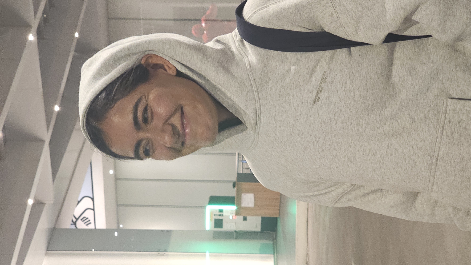 A woman in a gray hoodie with a dark blue backpack strap visible at the top of her shoulder is smiling at the camera. The hoodie appears to be branded with the words "ABERCROMBIE AND FITCH NEW YORK CITY" but these words are difficult to fully read. The background includes a light, modern architectural space with white structural beams overhead. A machine with green lighting and a counter with wood paneling can be seen in the mid-ground. The location is in the city of Puebla, Mexico.