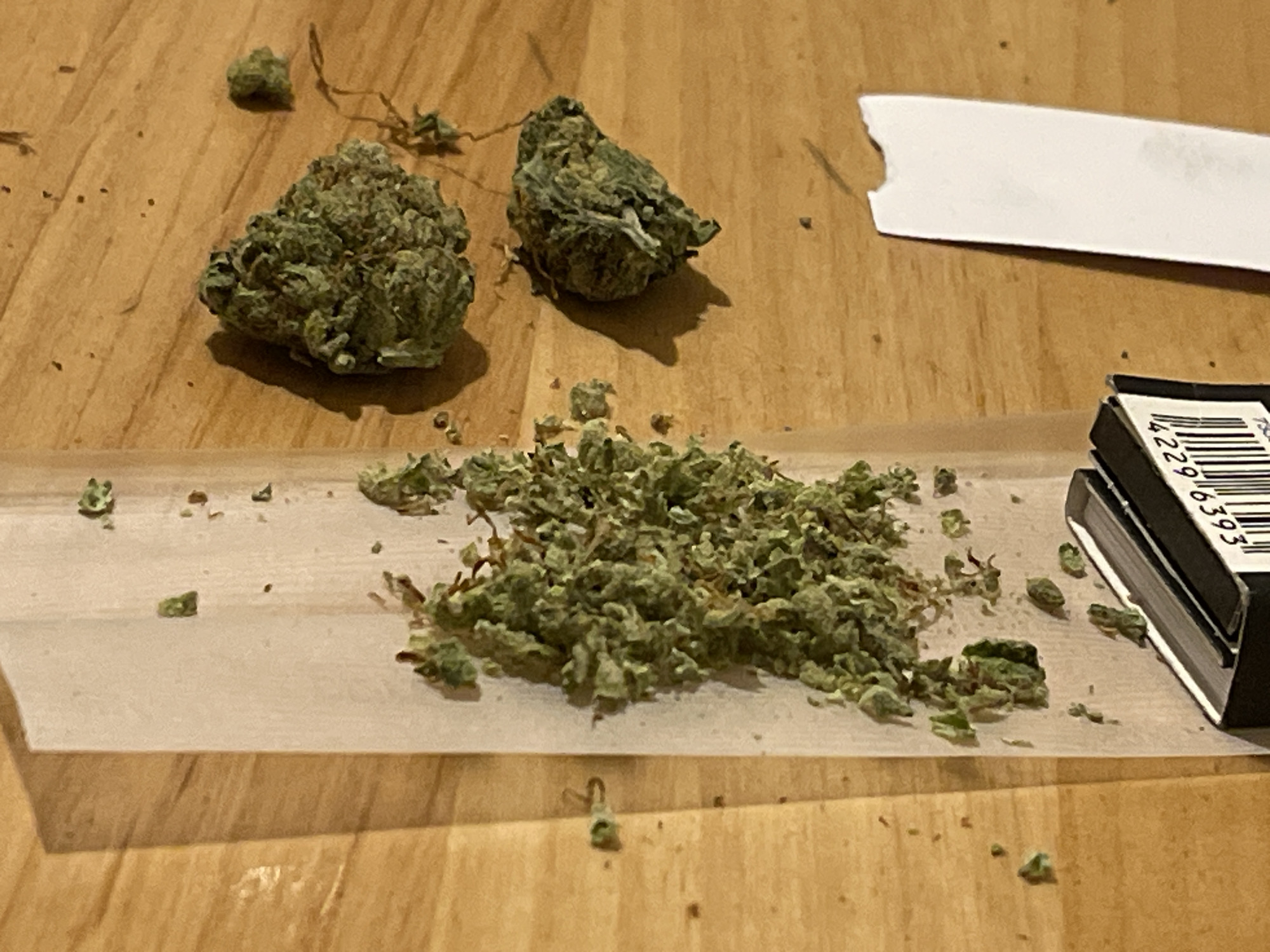 The image displays a static scene on a light brown wooden surface, possibly a tabletop, in the city of Unknown, Unknown. In the upper half of the frame, two distinct, intact cannabis buds are present. The bud on the left is compact and dark green, while the bud on the right appears slightly looser with visible lighter green and white fibrous material, casting a pronounced shadow. In the lower-middle section, a significant pile of finely ground green plant material, consistent with cannabis, is spread across an unfolded, light tan-colored rolling paper. This rolling paper extends across the lower portion of the frame. To the immediate right of the ground cannabis, resting partly on the rolling paper, is a small, black object resembling a pack of rolling papers or a matchbook, displaying a white strip with a black barcode marked with the numbers "42296393". Numerous small, dark green fragments of plant matter and scattered dark speckles are visible across the wooden surface, particularly around the cannabis buds and the rolling paper, indicating prior processing or handling. In the upper right quadrant, an irregularly torn piece of white paper or thin cardboard is also visible.