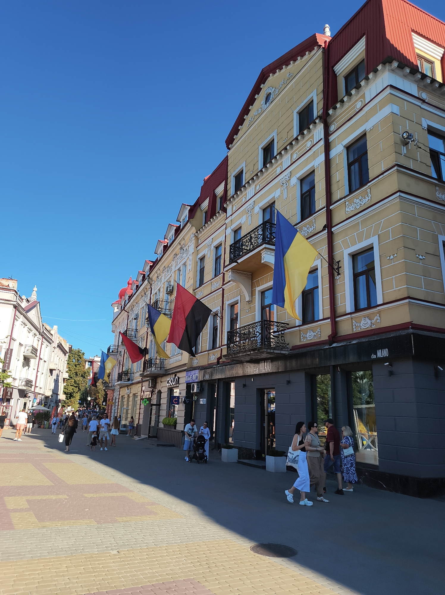 This image depicts a sunny day on Ternopil Street in Ternopil, Ukraine. The scene is dominated by a yellow building with a red roof, adorned with decorative elements and balconies from which Ukrainian flags and other national flags are hanging. The street is paved with colorful interlocking blocks and asphalt. Several people are walking along the street, some pushing strollers or carrying bags, and others engaged in conversation. Shops with large windows are visible along the building's facade, with some signage indicating businesses. The clear blue sky suggests pleasant weather.