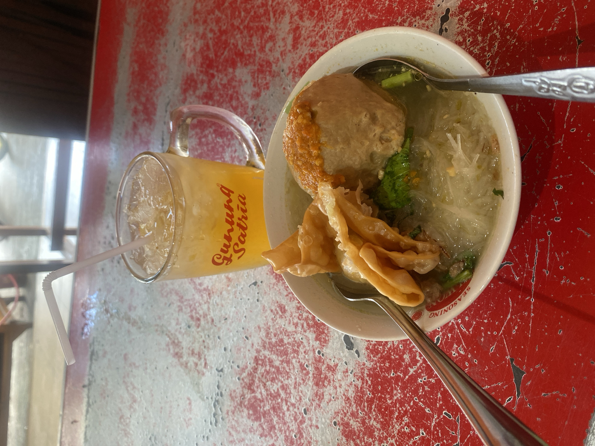 A close-up, eye-level shot shows a bowl of bakso (Indonesian meatball soup) and a glass of iced tea with a straw, placed on a textured red table. The bakso soup contains a large meatball topped with chili sauce, a few fried wontons, green vegetables, and thin rice noodles in a clear broth. The glass of iced tea is filled with ice cubes and has a bent white straw. The table has a distressed red painted surface with visible scratches and wear. In the background, blurry wooden furniture and a brightly lit outdoor area suggest a restaurant or eatery setting. The overall impression is of a casual meal enjoyed at an establishment, possibly in Sungai Sipai, Indonesia, given the "Gunung Satria" branding on the glass, which is a known business in the area.