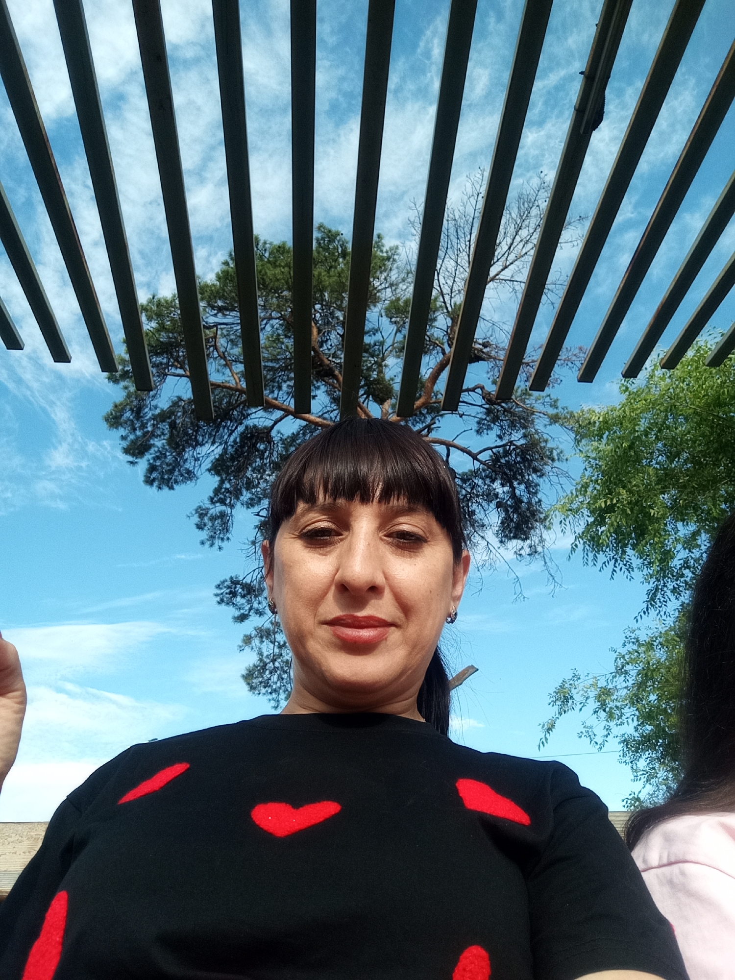 A woman, appearing to be in her late 30s or early 40s, is captured in a selfie taken outdoors. She has dark brown hair with bangs and is wearing a black t-shirt adorned with red heart-shaped patterns. A hint of another person, possibly female, wearing a pink top, is visible to the right of the frame. The background is dominated by a bright blue sky with wispy white clouds. Overhead, a wooden structure with parallel slats creates a visual framing effect. The foliage of trees, including a pine tree, is visible through the gaps in the slats. The setting appears to be a park or recreational area, possibly in the city of Belogorsk, Russia, given the context provided. The lighting suggests it is daytime, with the sun providing natural illumination. The overall scene is casual and relaxed.