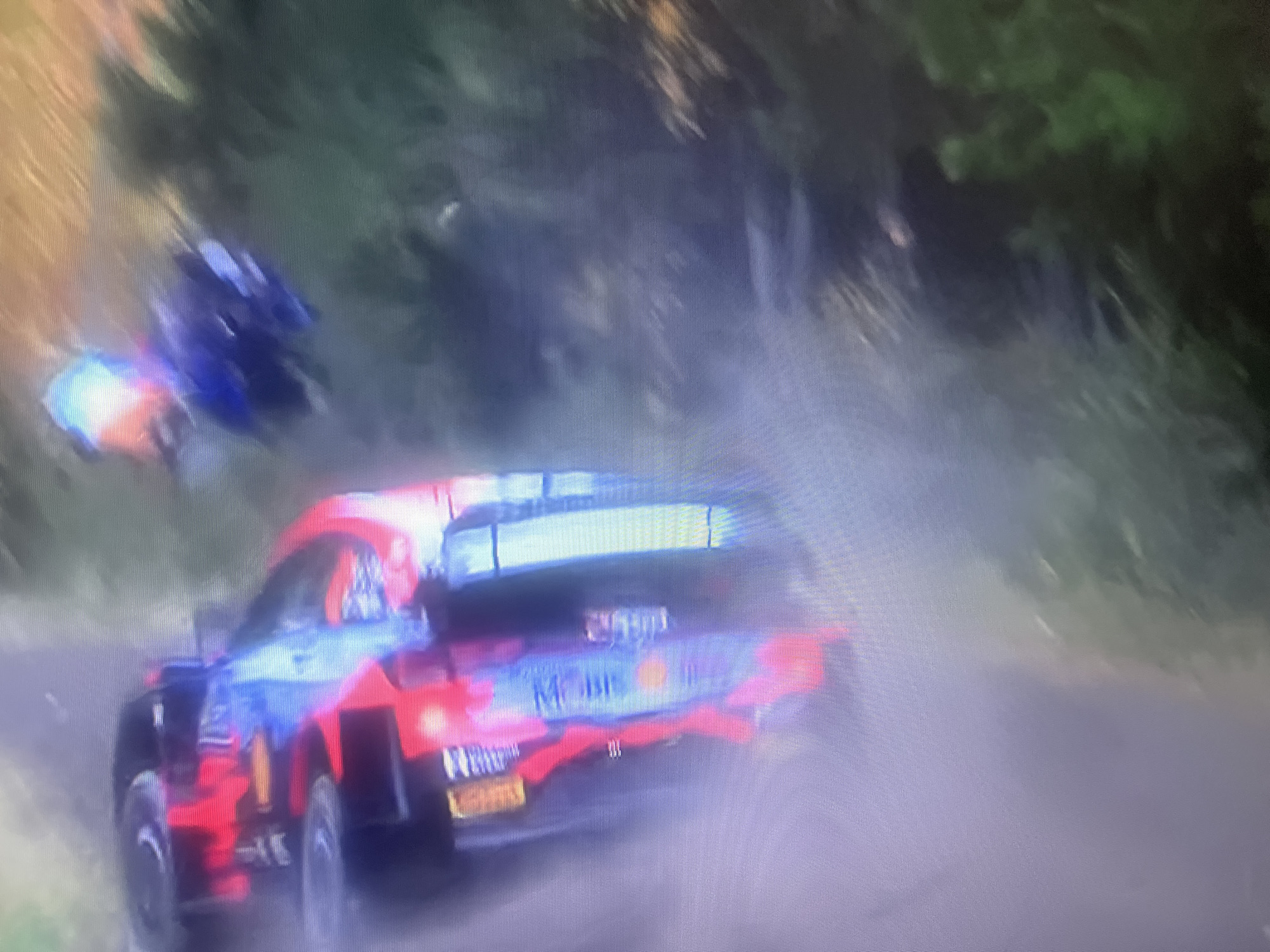 The image shows a red and blue rally car in motion, kicking up dust on a dirt road. The car appears to be a Hyundai i20 WRC. The background consists of trees and foliage, suggesting a natural or semi-natural environment. The lighting is somewhat harsh and appears to be from a daylight setting. The image is of low resolution and appears to be a screenshot or a photograph of a screen, possibly from a video game or a broadcast. There is no visible indication that this scene is located in the city of Masazir, Azerbaijan.