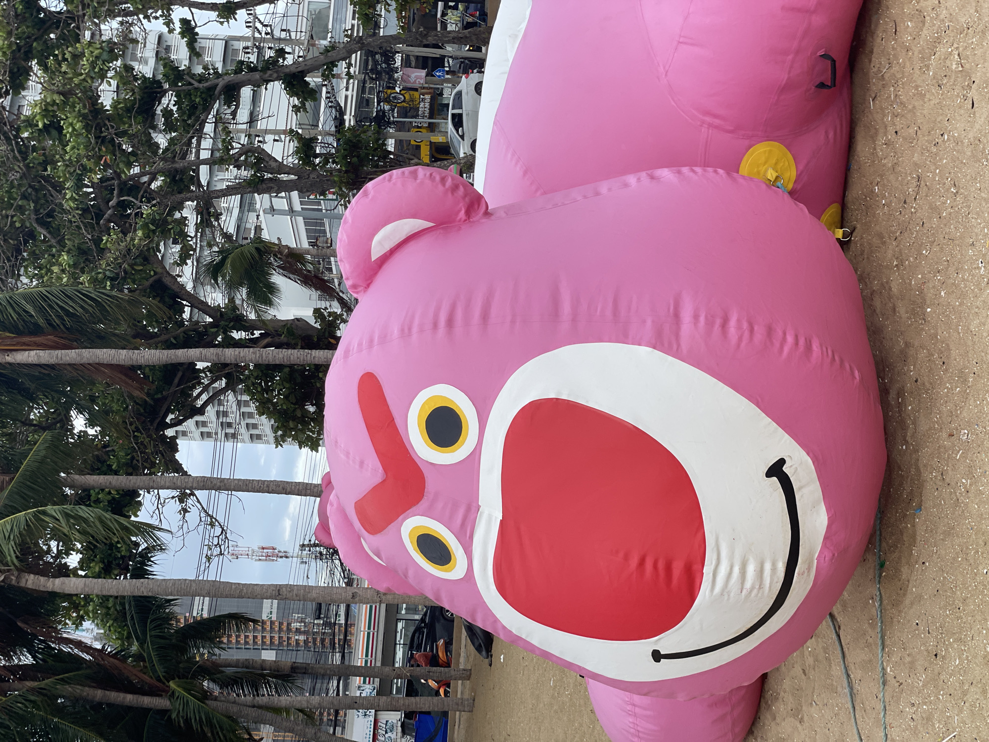 An inflatable, pink Lotso Huggin' Bear character from Toy Story is lying on a sandy beach in Pattaya City, Thailand. The bear is positioned in the foreground, taking up most of the frame. It is facing towards the right, with its large head and ears visible. The bear's face features angry red eyebrows, yellow eyes with black pupils, and a large red snout with a white outline. A smiling black mouth is also present. The inflatable is made of smooth, pink material with visible seams. In the background, palm trees and other foliage are visible, with urban buildings and a street with cars and a motorbike further in the distance. The sky is overcast. The beach appears to be a public area, possibly a tourist destination.