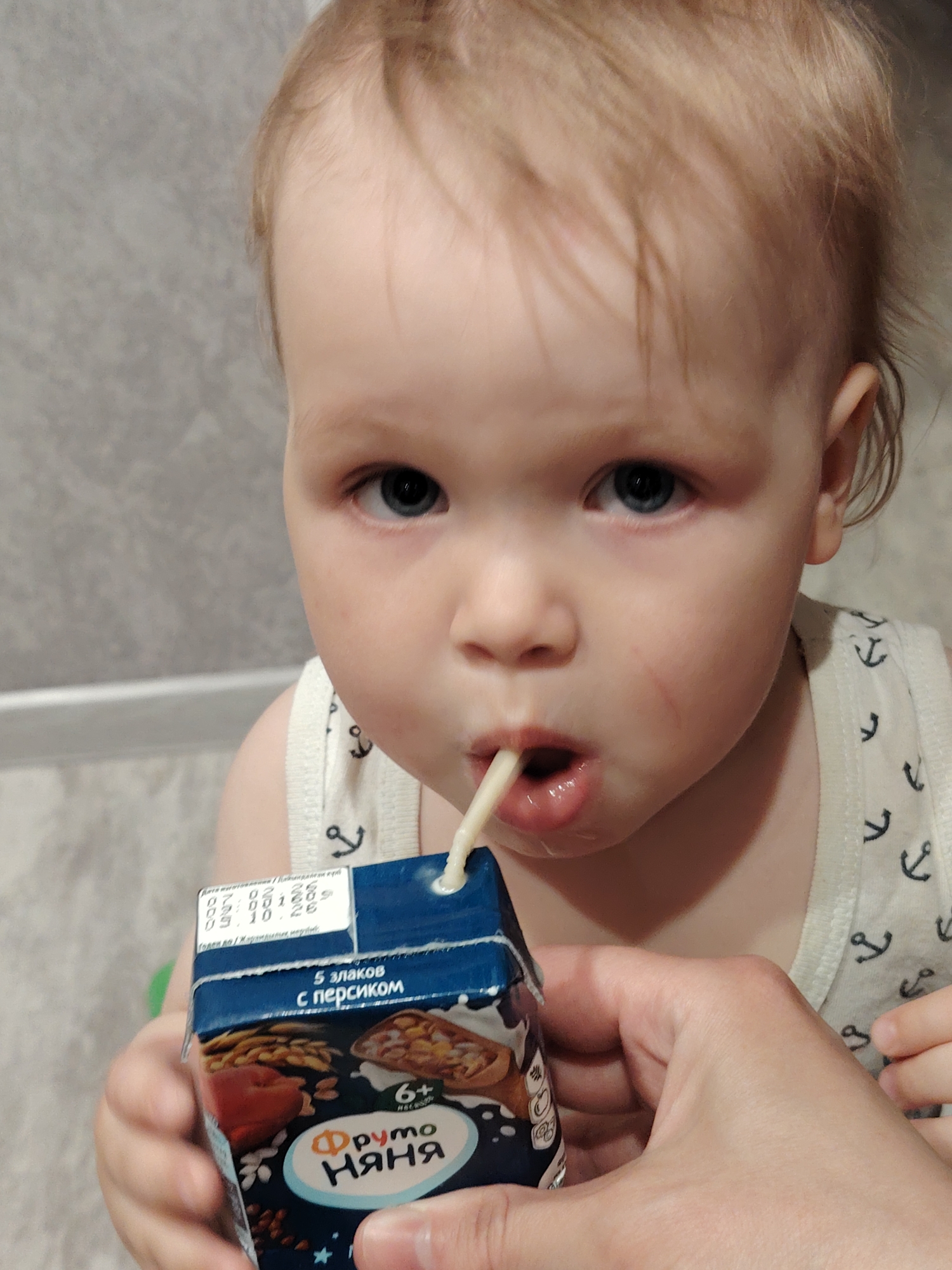 A toddler is drinking from a juice box using a straw. The box is blue and has a picture of fruit and the words "Фруто Няня" written on it. The juice box is being held by an adult hand. The toddler is wearing a white tank top with anchor patterns. The background appears to be a wall with a textured pattern. The juice box has an expiration date code on top, suggesting a product available for sale in Russia, though location of the exact photograph isn't provided. The juice box label is in Russian. Specifically, the juice box is "Фруто Няня" which is a popular brand of baby food and juices in Russia. This implies that the photo was taken in Russia or at least features products from that country. The toddler is drinking juice from the brand "Fruto Nyanya", this may be related to the Chaikovsky region in Russia.