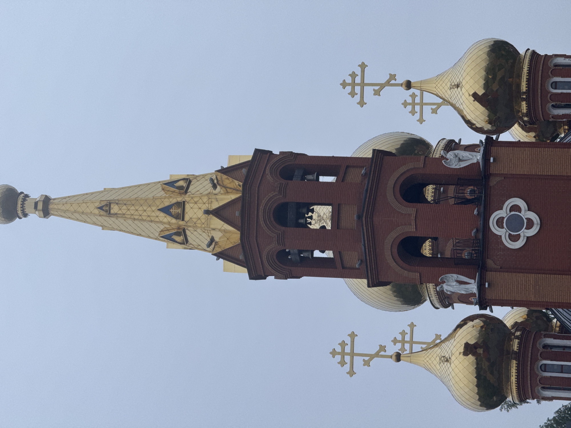 The image displays the exterior of a large, ornate Eastern Orthodox church or cathedral, viewed from a low angle against a clear, pale gray sky, suggesting an overcast day. The building is constructed primarily of red brick with intricate detailing.
Dominating the center is a very tall, slender spire, tapering to a point and covered entirely in golden, scale-like plating. Below it is a bell tower section, also made of red brick, featuring multiple arched openings that reveal bells hanging within.
Flanking the central structure are several golden, onion-shaped domes, characteristic of Orthodox Christian architecture. Each dome is topped with an elaborate golden Orthodox cross. One of the larger domes has a band of text at its base.
Notable details include white statues of angels placed on ledges of the brick facade, particularly visible near the base of the domes. A decorative white circular element with internal divisions is also visible on a red brick wall section. Small fragments of green foliage are barely visible at the bottom edge of the frame.
The visible text, in Cyrillic script, is primarily found on the golden band at the base of the large golden dome, clearly reading "СВЯТИТЕЛЬ НИКОЛАЙ ЧУДОТВОРЕЦ" (Saint Nicholas the Wonderworker). Further Cyrillic inscriptions, appearing on gold-colored surfaces, are also present inside the arched openings of the bell tower, though they are less distinctly legible, hinting at religious names like "АРХАНГЕЛ" (Archangel).
The architectural style strongly indicates the location as an Orthodox Christian country. Given the context, this is likely the St. Nicholas Cathedral in Chaikovsky, Russia.
