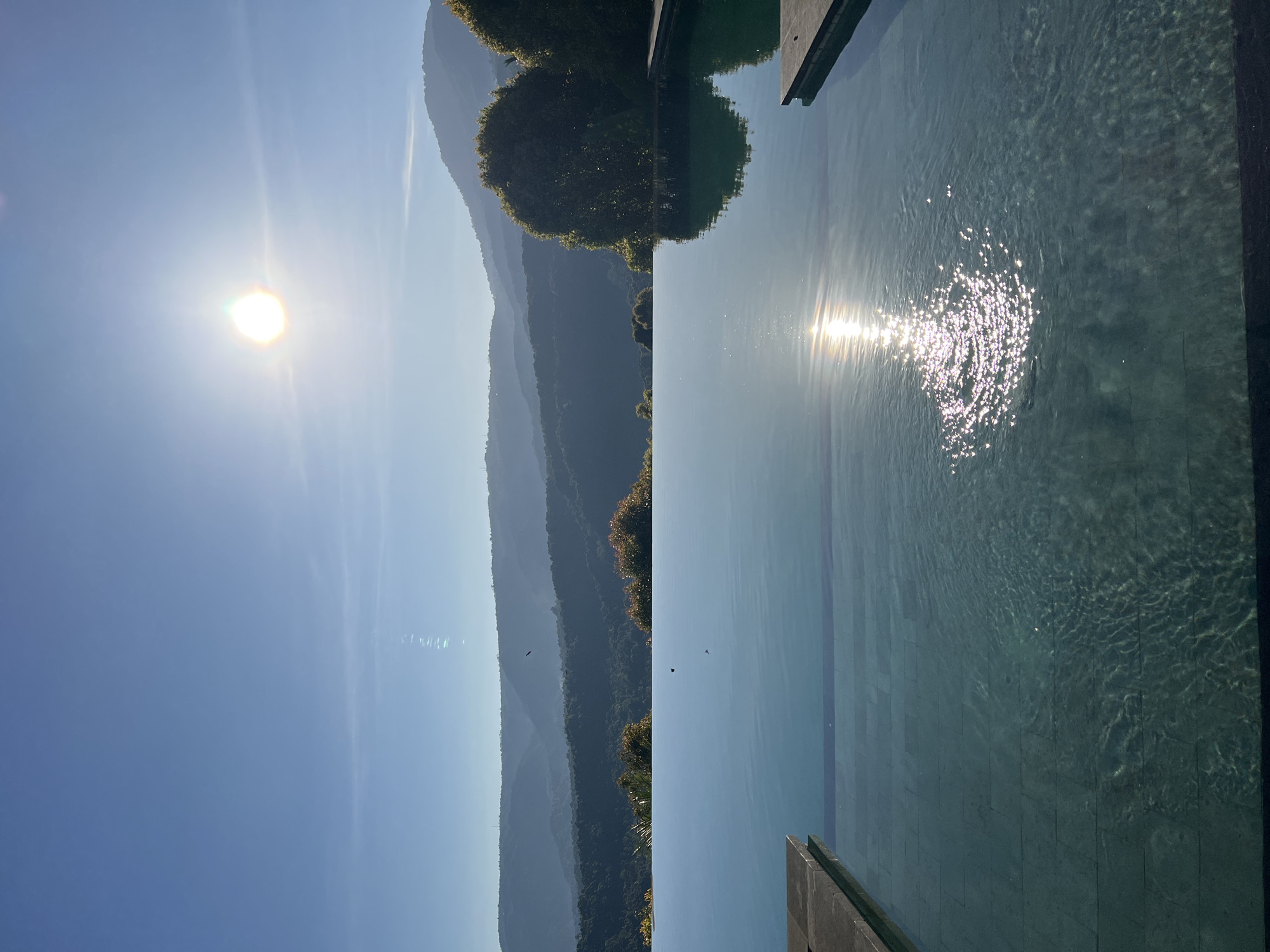 A breathtaking view of an infinity pool overlooking a verdant valley under a bright, sunny sky. The sun, a brilliant orb, shines intensely in the upper right quadrant, casting shimmering reflections on the pool's surface. The water, clear and inviting, reveals the tiled bottom of the pool. In the distance, rolling hills covered in dense green foliage stretch towards the horizon, their contours softened by a hazy atmosphere. The scene suggests a tranquil, luxurious retreat, likely a resort or private villa, in the serene landscape of Busungbiu, Indonesia.