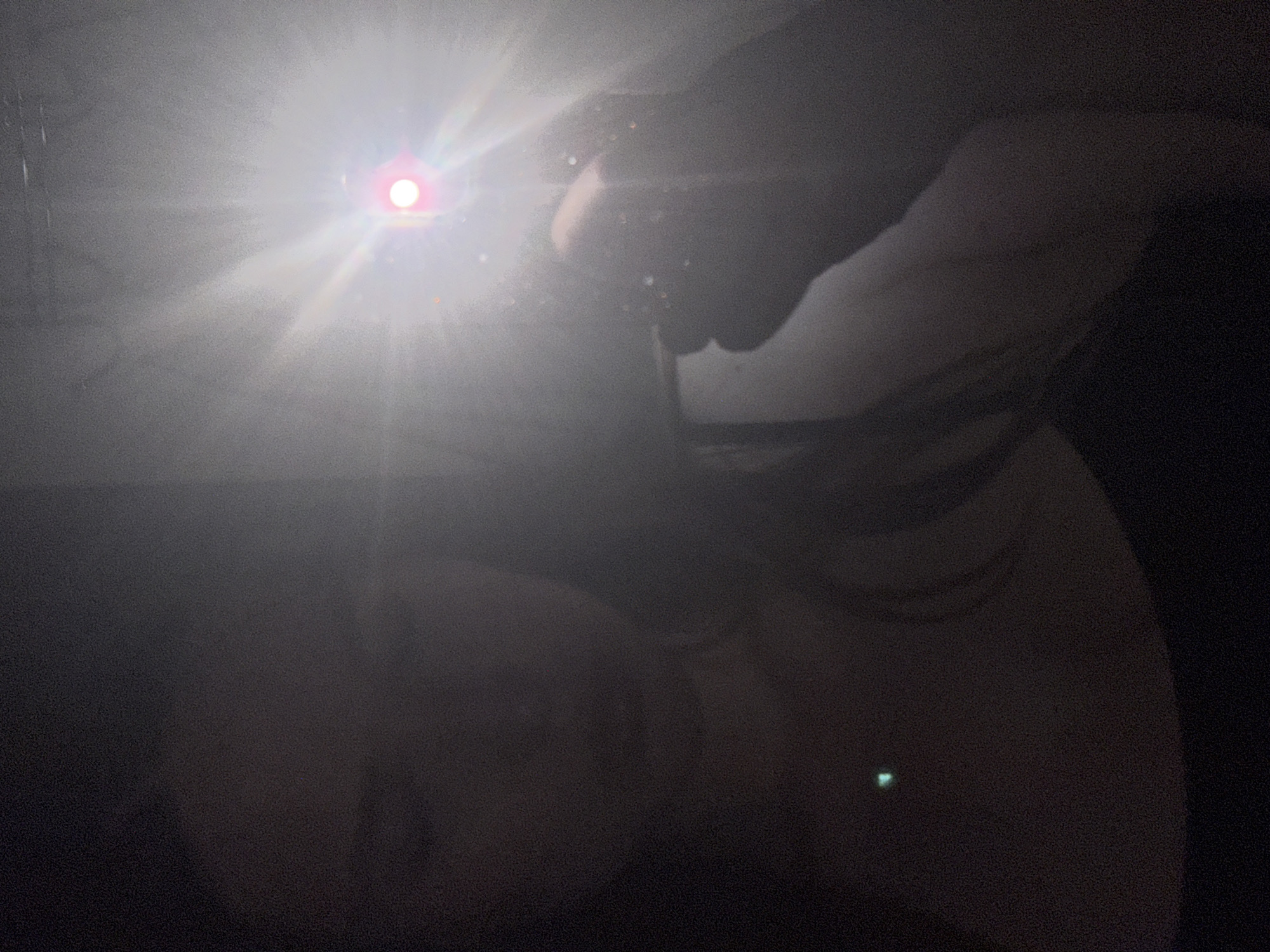 The image is a dimly lit selfie taken by a person holding a light source. The person's face is largely obscured by shadow and the glare of the light, but features such as the nose and mouth are discernible. A bright red light source, possibly a camera flash or a small light, is visible to the right of the frame, creating lens flare and illuminating a hand holding the light. A small green light is also visible on the person's chest. The background is dark and indistinct, with some lighter areas suggesting a reflective surface or background. Without additional information or context, the specific location in Chaikovsky, Russia cannot be determined. The image captures a personal moment, likely taken indoors or in a low-light environment, with the focus on the interaction between the person and the light source.