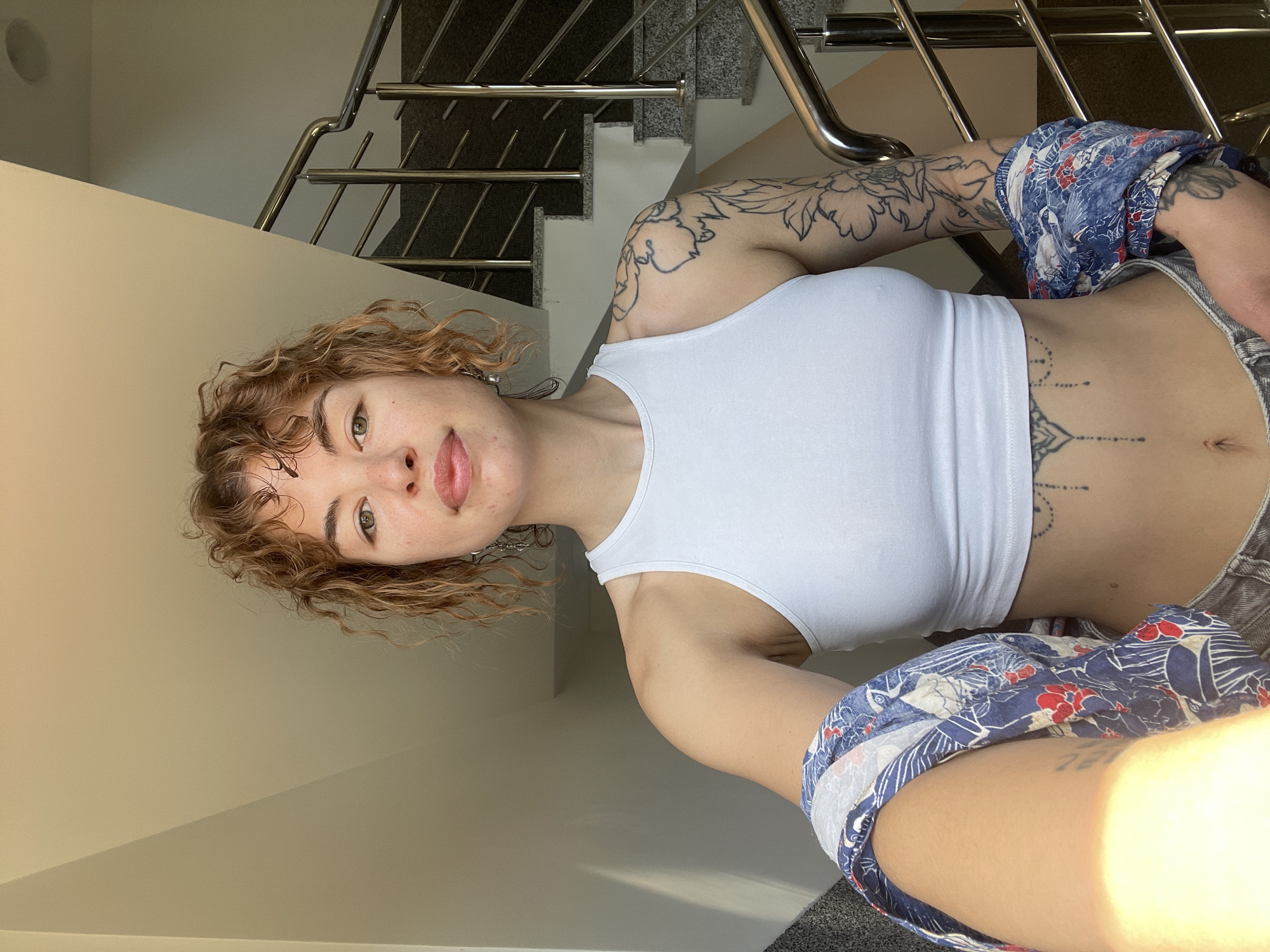 A woman with light skin and curly brown hair is positioned in front of a staircase with a metal railing. She is wearing a white crop top and patterned blue and white shorts. The woman has tattoos on her arms and abdomen. The background shows stairs and a white wall. The image appears to be taken indoors, possibly in a residential building, though the specific location in Boreti, Montenegro, is not discernable from the image. There is no other person, subject, object, action, interaction, or other specific environment element presented in the image.