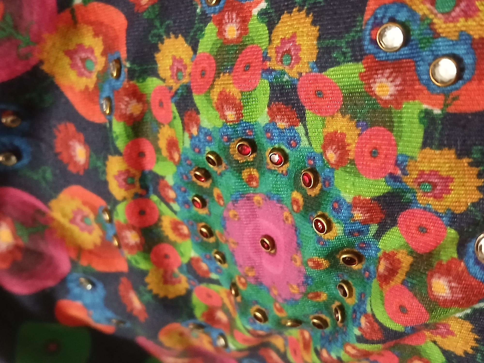 The image shows a close-up of fabric with a colorful floral pattern. The pattern includes various flowers in shades of red, orange, yellow, green, blue, and pink, set against a dark blue background. The fabric is also adorned with metallic studs, some of which appear to have red or white jewels or reflective material set in them. The studs are arranged in a radial pattern in some areas. The image does not provide any information about the location in the city of Bengaluru, India, people, actions, interactions, or the environment.