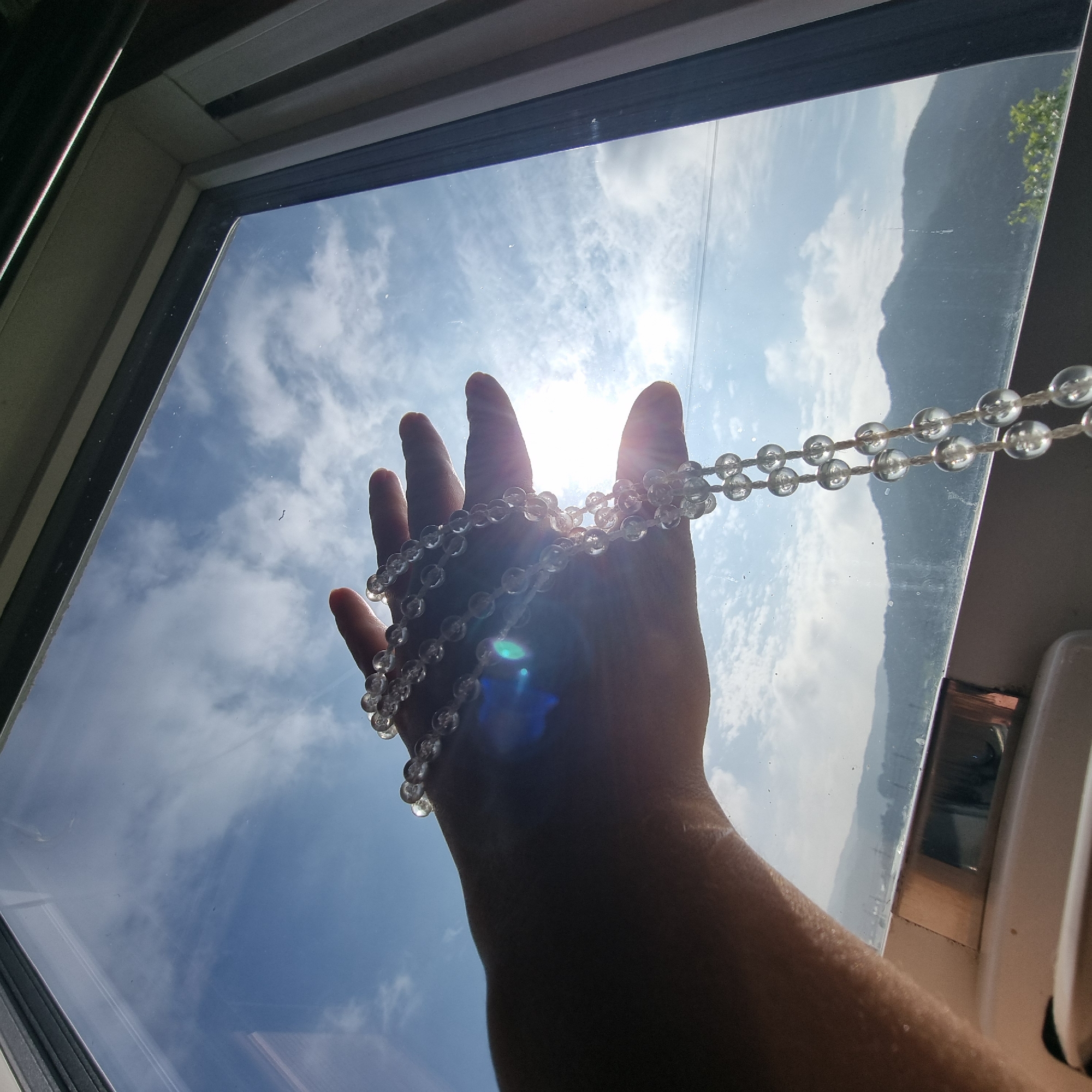 An over-the-shoulder, close-up shot shows a hand holding a string of translucent, spherical beads against a bright sky seen through a window. The hand is silhouetted, with the sun creating a lens flare and light rays that pass through the beads, illuminating them. The sky is blue with scattered white clouds, and a bright sun is visible. In the background, seen through the window, are hazy green mountains and a few trees. The scene appears to be indoors, with the window frame visible in the foreground. The image may have been taken in Dunnae-myeon, South Korea, given the mountainous terrain and architecture visible.