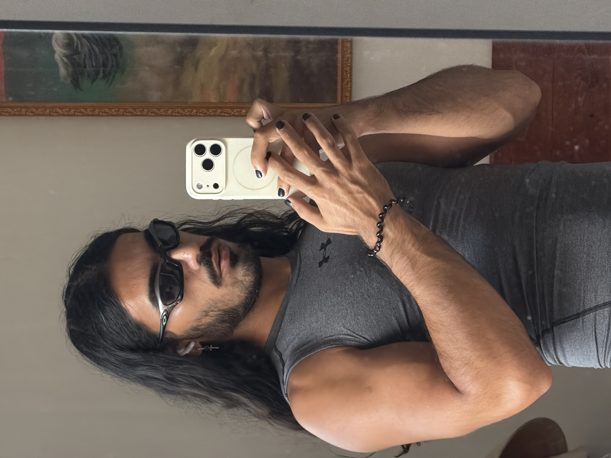 A young man with long, dark hair, sunglasses, and a grey sleeveless shirt is taking a mirror selfie in North Kuta, Indonesia. He is holding a white smartphone in his hands, which are adorned with black nail polish. A silver cross earring is visible in his left ear, and a beaded bracelet is on his right wrist. Behind him, a framed painting hangs on the wall. The setting appears to be an indoor space, possibly a bedroom or bathroom, with soft lighting.
