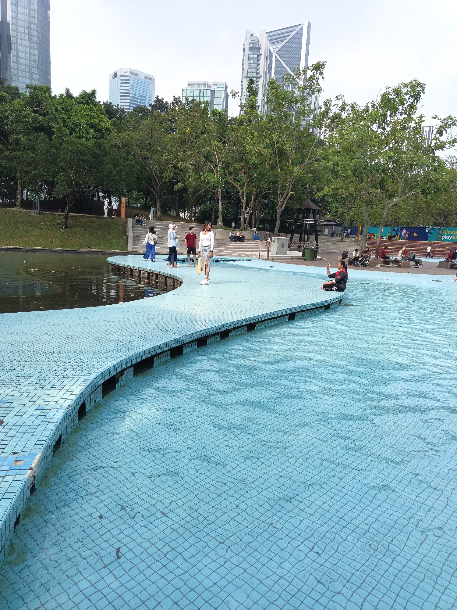 This is an outdoor image taken at the Kuala Lumpur City Centre Park (KLCC Park) in Kuala Lumpur, Malaysia. The foreground features a blue tiled, shallow pool with a curved, raised walkway made of similar blue tiles. Several people are visible on the walkway and surrounding areas. In the midground, a group of four individuals, two women in white and blue clothing and two men in maroon and white shirts, are standing on the walkway near the water's edge. Another individual, a woman in a white top and grey skirt, is standing further along the walkway holding a yellow object. To the right, a person is seated on the edge of the walkway, taking a photo with a smartphone. In the background, a body of water, possibly a lake or pond, is visible to the left of the walkway. Beyond that, a lush green park area with trees leads up to a backdrop of modern skyscrapers, characteristic of the Kuala Lumpur city skyline. A small, open-air pavilion structure with dark roof and pillars is situated within the park. Further in the background, there are more people relaxing on benches and stairs, and children playing in a playground area with blue and yellow structures. The sky is overcast.