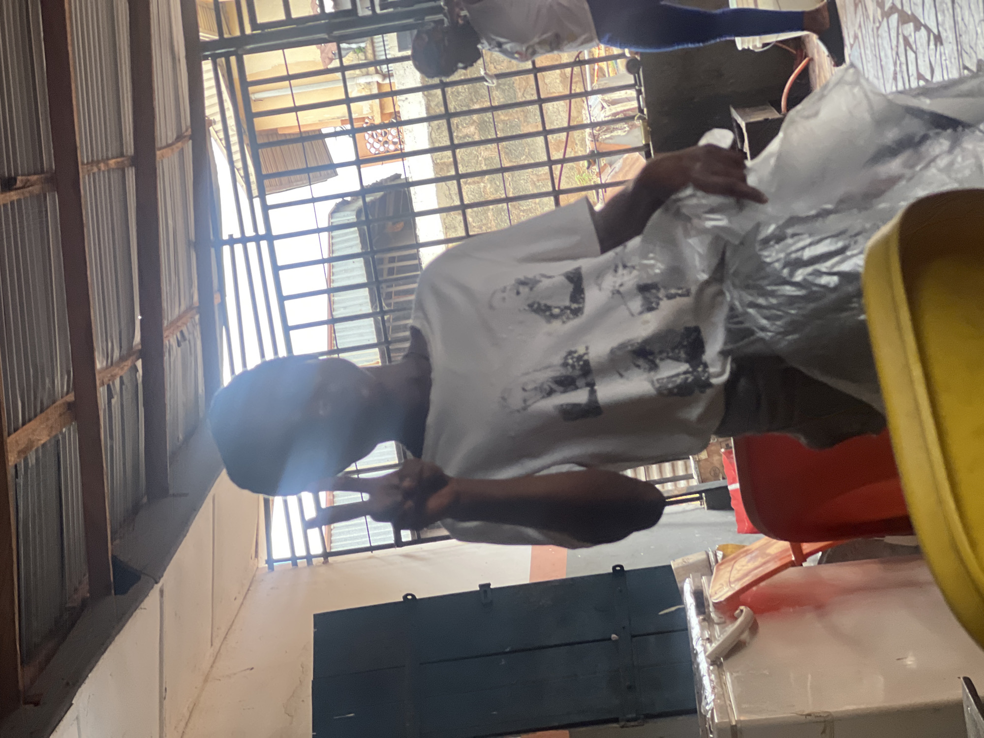 In Nkpor, Nigeria, a person wearing a white t-shirt with images on it is posing with a peace sign, holding a plastic bag. The person appears to be standing in an outdoor or semi-outdoor area with a corrugated metal roof supported by wooden beams. Behind the person, a metal gate or bars are visible, with buildings and more corrugated metal structures in the background. To the left of the person, a dark blue door or cabinet is present. A second person, wearing a white t-shirt and blue pants, is visible further back in the scene, partially obscured by the gate. The environment appears to be somewhat rustic, with visible construction elements and a sense of casual outdoor activity.