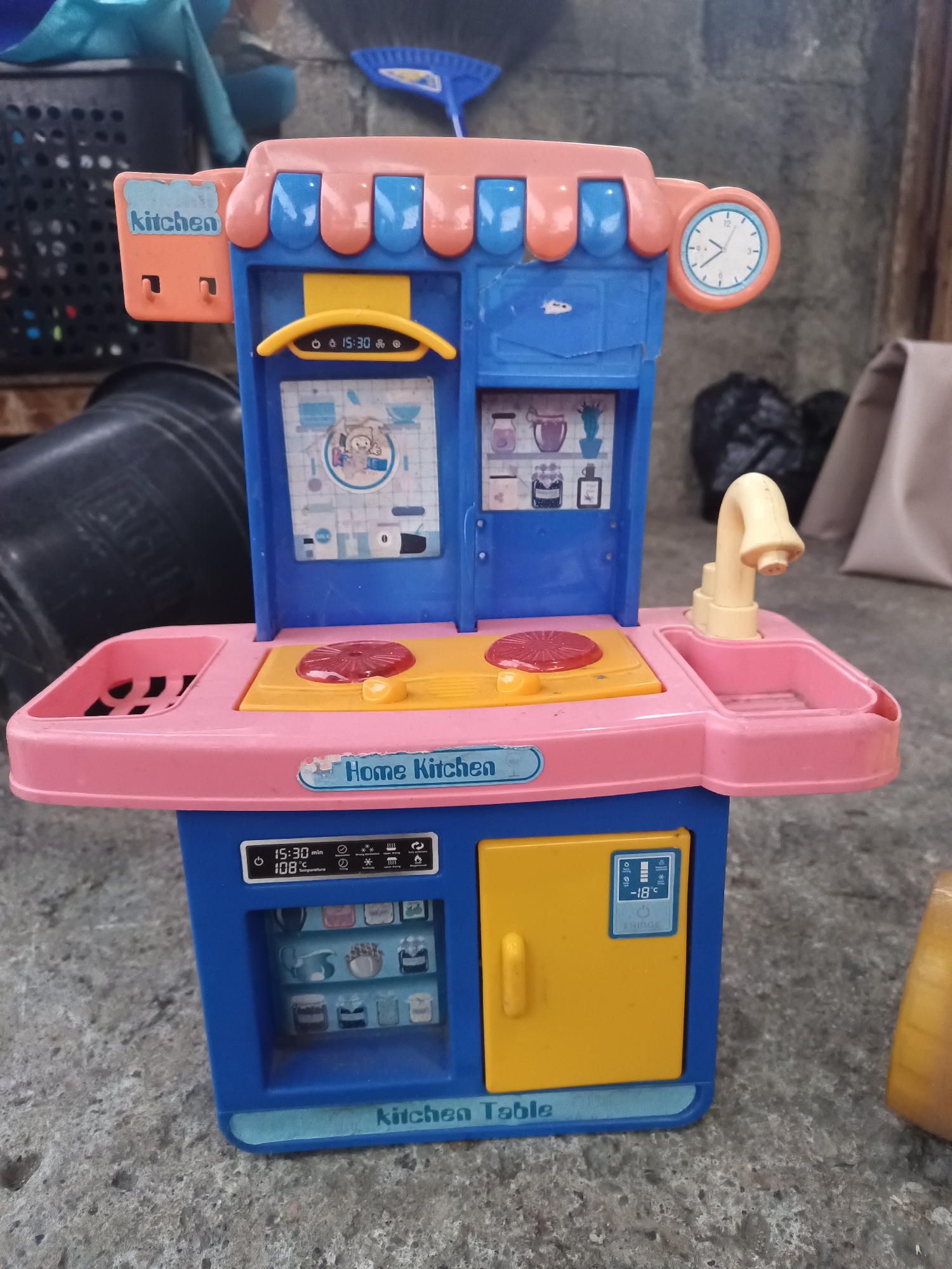 A toy kitchen playset, predominantly pink and blue with orange accents, sits on a concrete surface in what appears to be an outdoor or semi-outdoor area. The playset features a stove with two red burners, a sink with a beige faucet, and an oven with a yellow door. Above the stove, a panel displays a clock and various icons. A label on the playset reads "Home Kitchen." To the left of the playset, a dark plastic bin and a blue object, possibly a broom, are visible. In the background, a textured wall and a rolled-up beige fabric are present. The lighting suggests daytime. The scene is consistent with a residential backyard or a similar outdoor space in South Jakarta, Indonesia. There are no people present in the image.