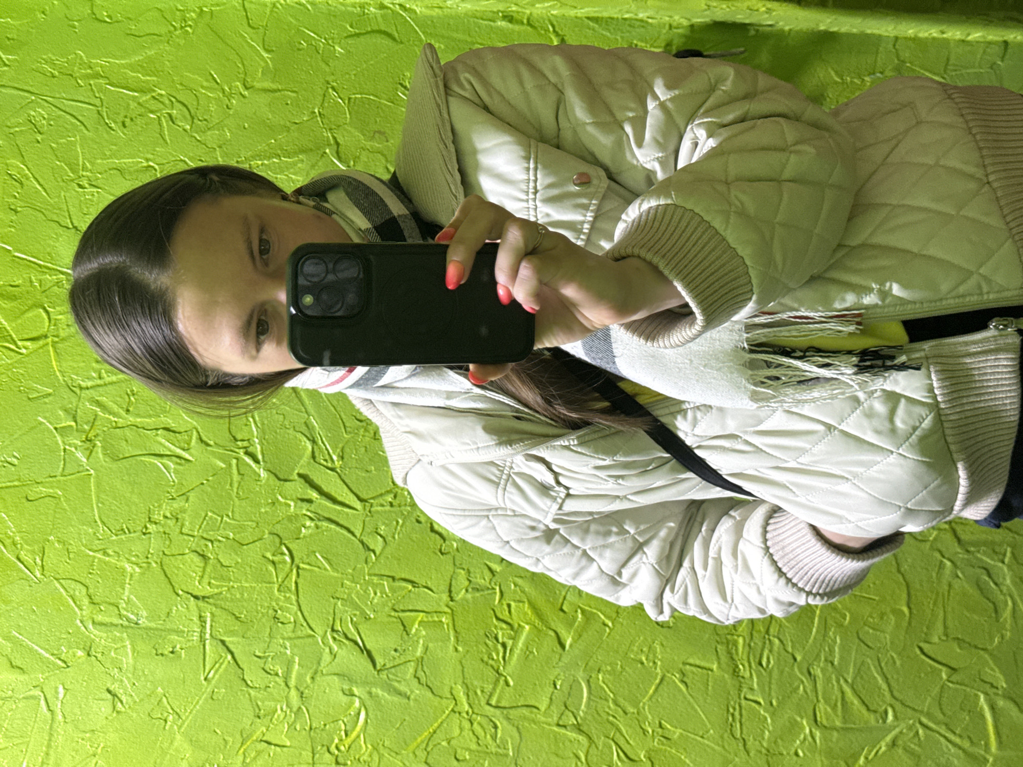The image captures a selfie taken by a person, presumably female, reflected in the screen of a dark-colored smartphone, likely an iPhone, which she holds up. Her face, with long dark hair parted in the middle, is partially visible in the phone's reflection. Her eyes are dark. She is wearing a light beige or off-white quilted jacket with ribbed cuffs and collar, layered over what appears to be a plaid or striped scarf. Her fingernails have a distinctive orange-to-red gradient manicure, and a ring is visible on her right ring finger. A black strap, possibly from a bag, is also visible. The background is a vibrant, textured lime green wall, suggesting an indoor setting or a specially designed space. The image is oriented sideways, with the person appearing on the right side against the green wall. No text is visible in the image. The photo was taken in Chaikovsky, Russia.