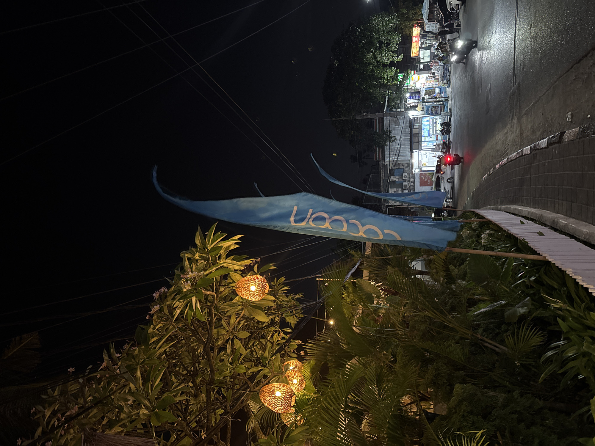 This image depicts a nighttime scene on a street in Kuta, Indonesia. A blue flag with the word "Cocoon" in white lettering is prominently displayed on the left side of the frame. To the left of the flag, lush green tropical foliage is illuminated by several woven, pendant lights, creating a warm, inviting atmosphere. A white picket fence runs along the edge of a sidewalk.
The street itself is paved and shows signs of activity. Several vehicles are visible, including cars and motorcycles, some of which have their headlights on, indicating movement. A red taillight of a motorcycle is particularly noticeable. In the background, buildings line the street, with shops and businesses displaying illuminated signs. The sky above is dark, with visible power lines crisscrossing the frame, typical of a city at night. The overall impression is a lively street scene in a tropical urban environment.