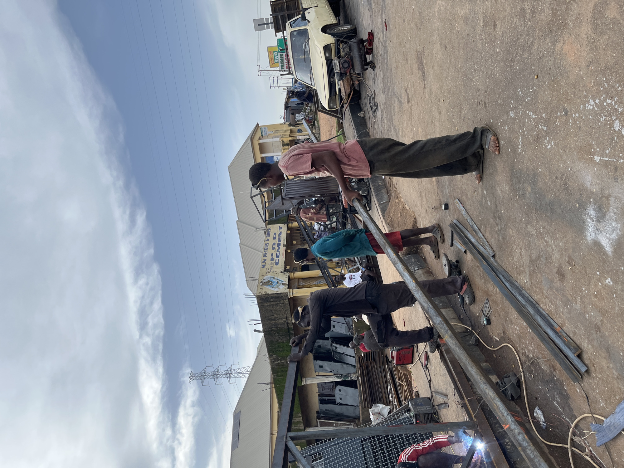 This image depicts an outdoor scene in Gura Top, Nigeria, likely during the daytime with a cloudy sky. The setting appears to be a roadside workshop or construction area. Several individuals are engaged in welding and metalworking activities.
In the foreground, a person wearing a pink shirt and dark pants is leaning over a metal structure, seemingly supervising or working on it. To the right, another person in dark pants and a dark long-sleeved shirt is also working on metal, with a welding torch actively producing sparks. Further back, a person in a blue shirt and another in a white t-shirt are visible amidst the ongoing work. A light-colored vehicle, possibly a car or utility vehicle, is parked in the background, with its engine exposed, suggesting it might be under repair or modification. Various metal rods and equipment, including a welding machine, are scattered around the work area.
A building with signage is visible behind the workers. The sign reads "M.N PETERS & SONS P.O.P CEMENT". The overall atmosphere suggests a busy work environment focused on metal fabrication or repair. The presence of a power pylon and overhead electrical lines in the sky indicates an urban or semi-urban setting.