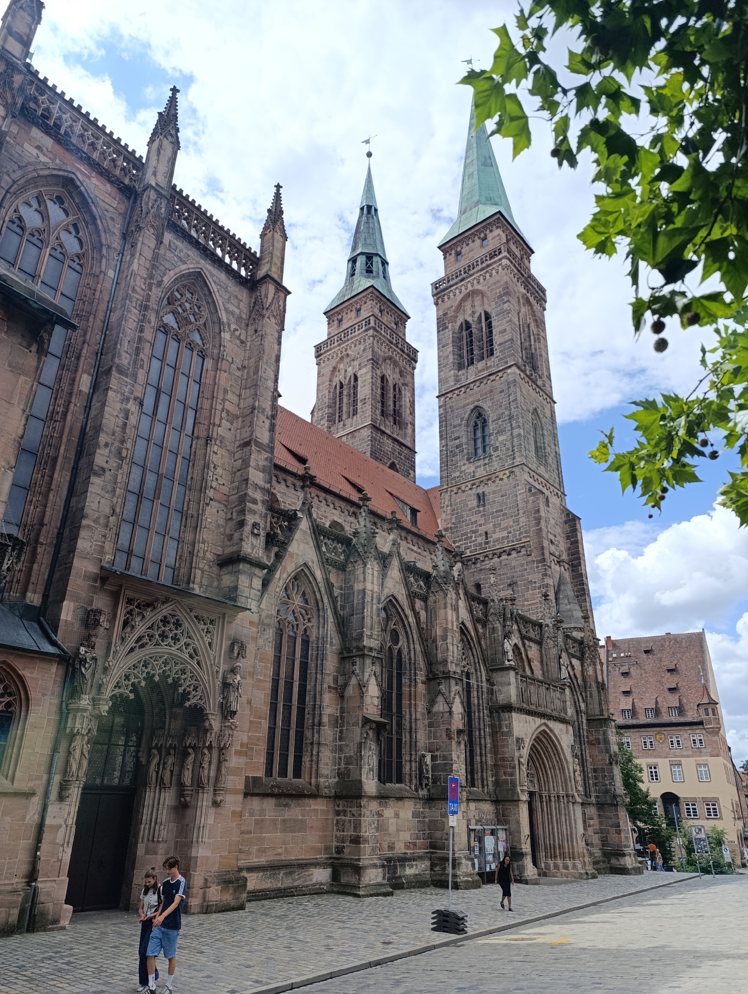 A long shot captures the exterior of St. Lorenz Church, a prominent Gothic cathedral in Nuremberg, Germany, under a cloudy sky. The church, constructed from stone, features two tall spires with green conical roofs, intricate architectural details, and large stained-glass windows. A cobblestone street runs in front of the building. Two young people, a male and a female, walk on the street near the entrance. In the background, a multistory building with a red tiled roof and several windows is visible, as well as lush green foliage from a tree in the foreground.