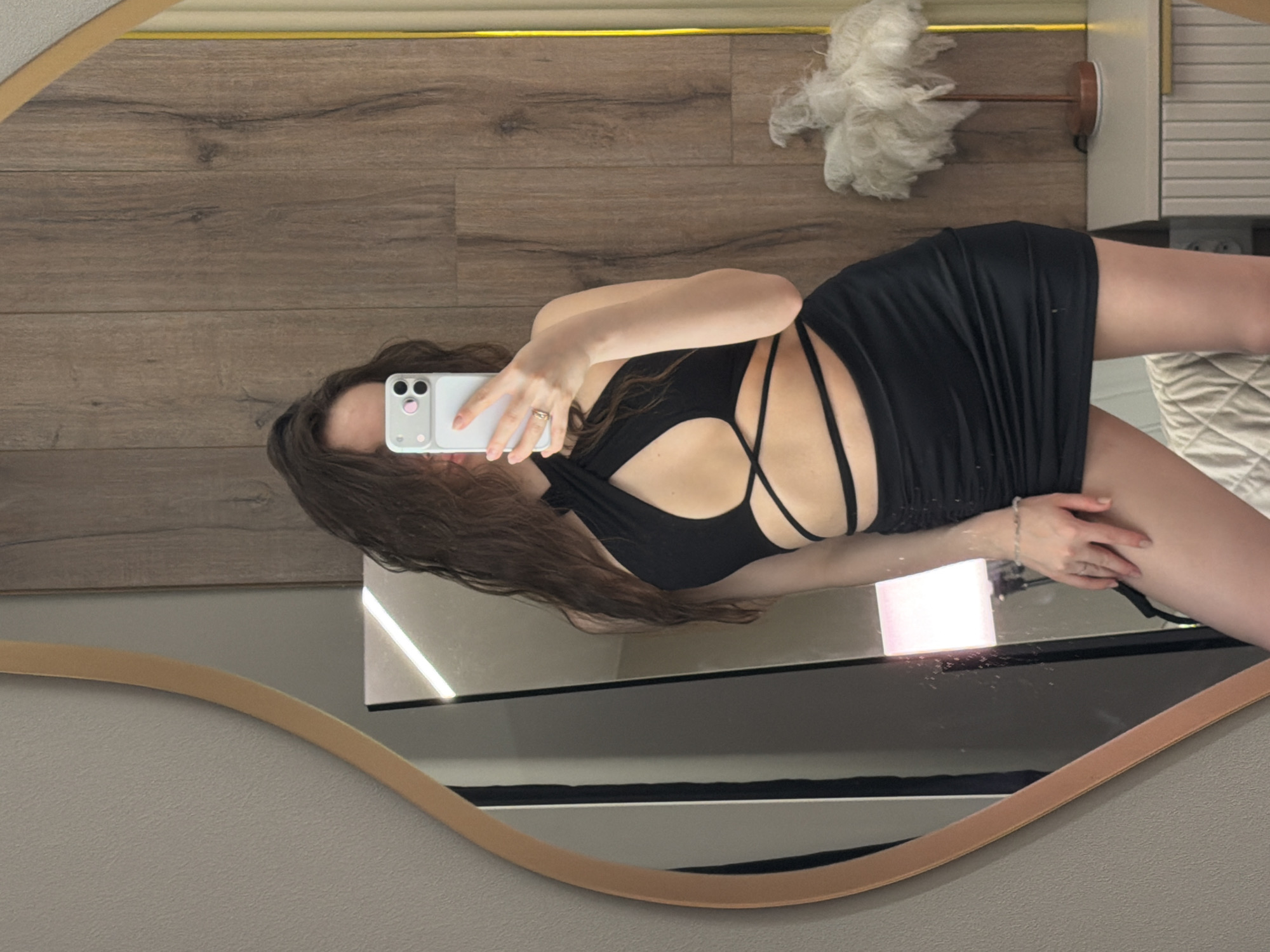 The image depicts a woman taking a selfie in a mirror. She is wearing a short black dress with cutouts in the chest area and straps. Her hair is long and wavy. She is holding a smartphone in her right hand, and her reflection is visible in the mirror. The background includes a wooden wall and what appears to be a decorative object hanging on the wall. There is no indication of the location being Chaikovsky, Russia, in the image.