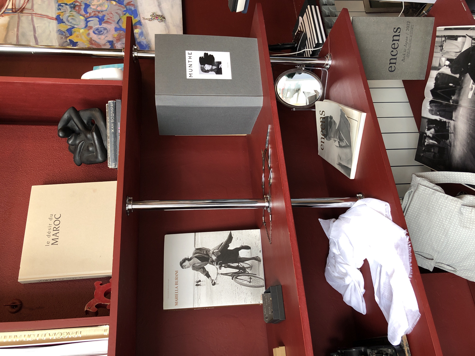 The image depicts a close-up view of a red shelf unit, likely inside a shop or store, possibly in Moscow, Russia, as suggested by the Cyrillic text on a book. The shelves contain various items, including books ("le désir du MAROC" by MARIELLA BURANI and "ЖАН БОДРИЙЯР"), a gray box labeled "MUNTHE", a small round mirror, a white scarf, and several other items. The lighting appears to be ambient, and the overall impression is of a cluttered, but organized shelf display.