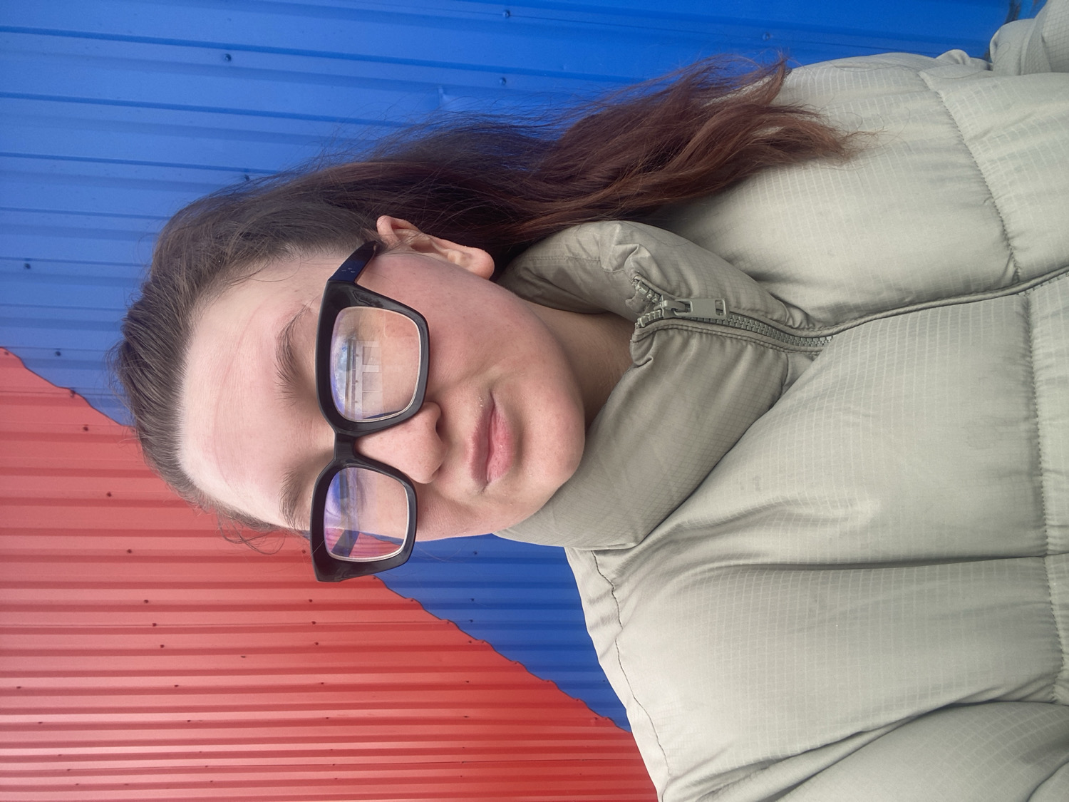 A close-up portrait shows a young individual, likely female, with fair skin and long, reddish-brown hair visible from the side. She wears dark-framed rectangular glasses, with reflections showing a bright, grid-like pattern on the lenses. Her expression is neutral to slightly pensive. She is dressed in a light-colored, possibly light green or beige, puffer jacket with a visible zipper and a subtle grid pattern on its fabric, suggesting cooler weather.
The background features a distinctive corrugated metal wall, diagonally split into two vibrant colors: red on the lower left and blue on the upper right. This wall appears to be the exterior cladding of a building. The person appears to be posing for the photograph in an outdoor setting, likely against this building in Chaikovsky, Russia. There is no visible text in the image.