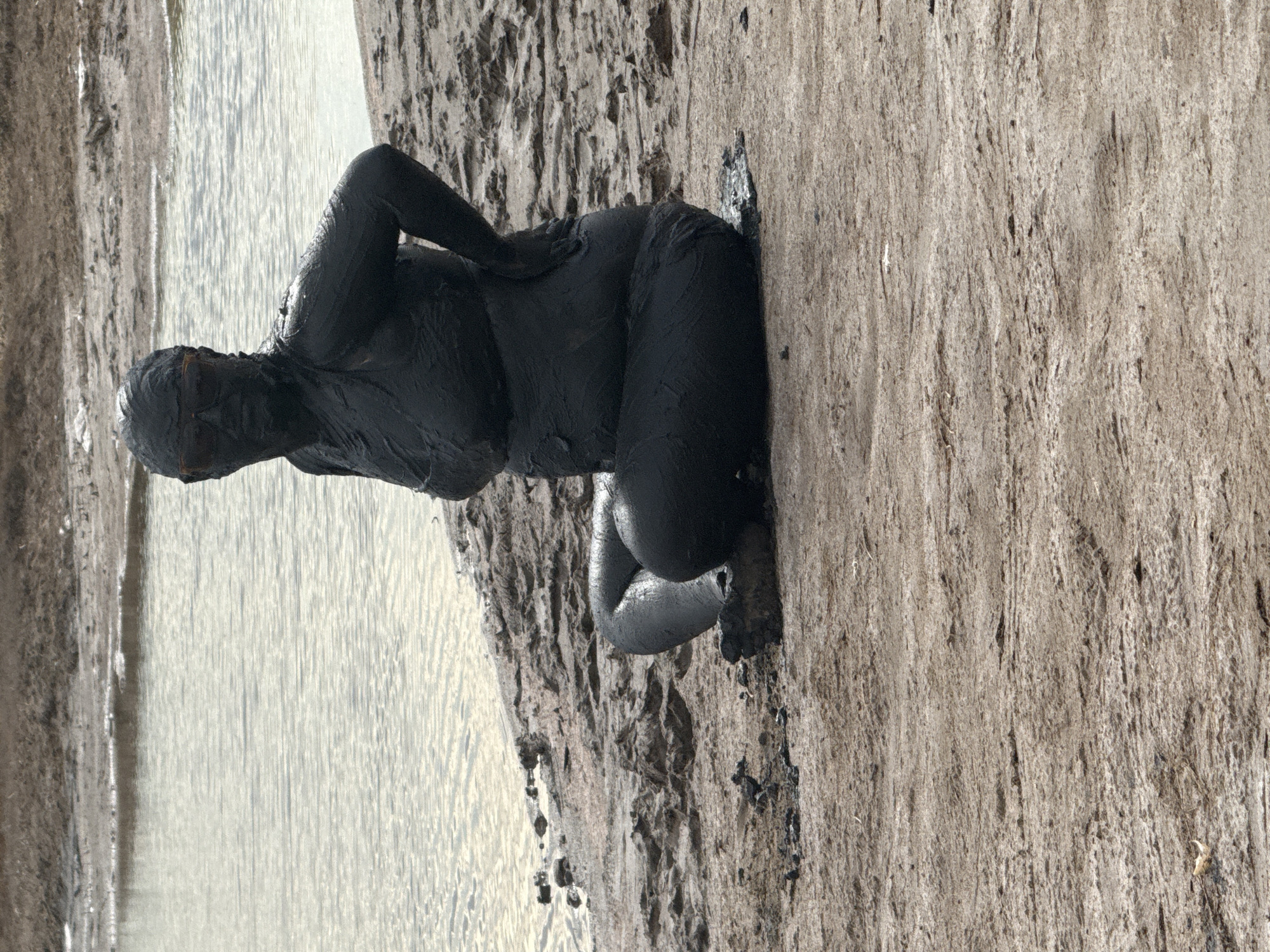 A person, covered head-to-toe in dark mud, sits on a muddy bank next to a body of water. The person is wearing sunglasses. The mud appears to be a thick, viscous substance. The surrounding environment is sandy and barren, with the water appearing murky. The scene is located in the Yamyyshevsk rural district of Kazakhstan.