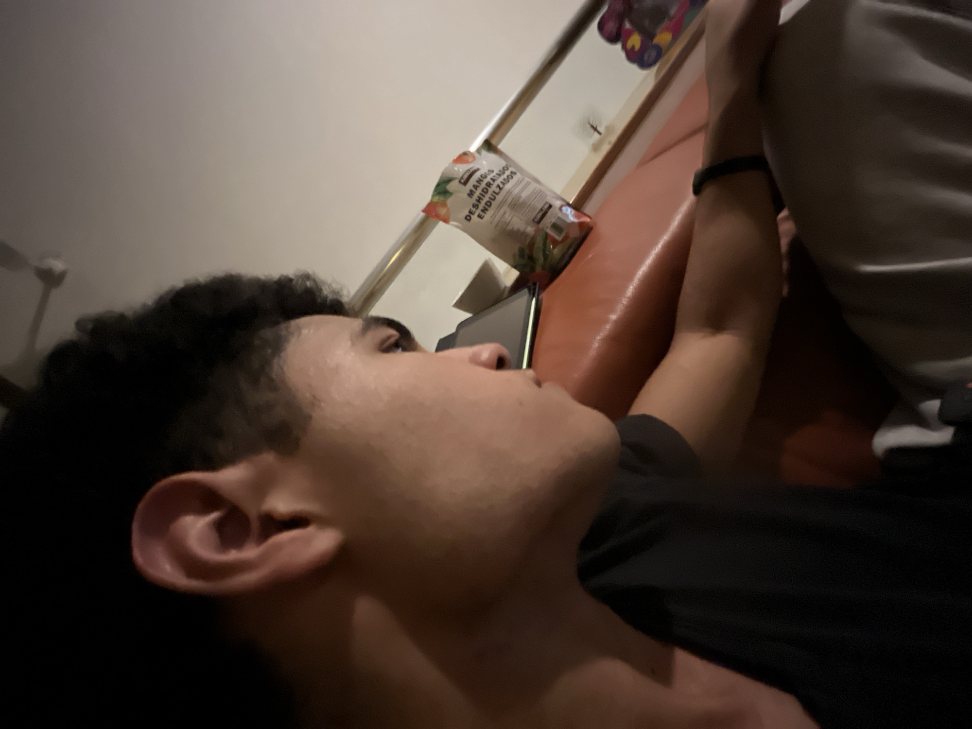 A young man is lying on an orange couch, facing away from the camera. He is wearing a black t-shirt and has dark curly hair. His right arm is visible and has a black fitness tracker on the wrist. A bag of "Mangos Deshidratados Endulzados" (Sweetened Dehydrated Mangoes) is on the couch near his head. A mobile phone is resting between the bag of mangoes and his face. The location is not specified in the image and there are no visual clues of being in the city of San Bernardino Tlaxcalancingo, Mexico.