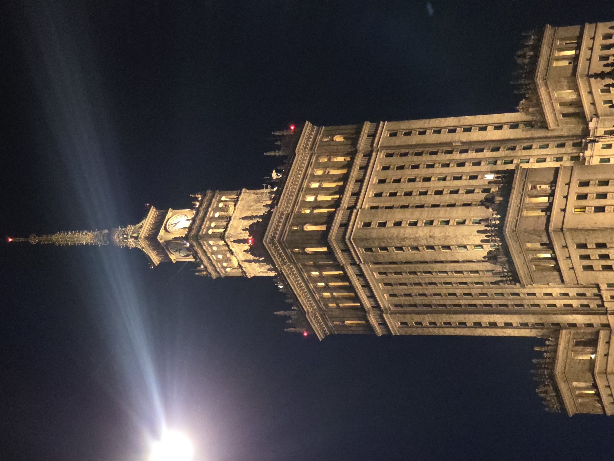 At night, a low-angle, rotated view shows the Palace of Culture and Science in Warsaw, Poland. The building is illuminated with yellow light against a dark sky. The structure's architectural details, including windows, spires, and the clock tower, are visible. A bright light source obscures the base of the building.