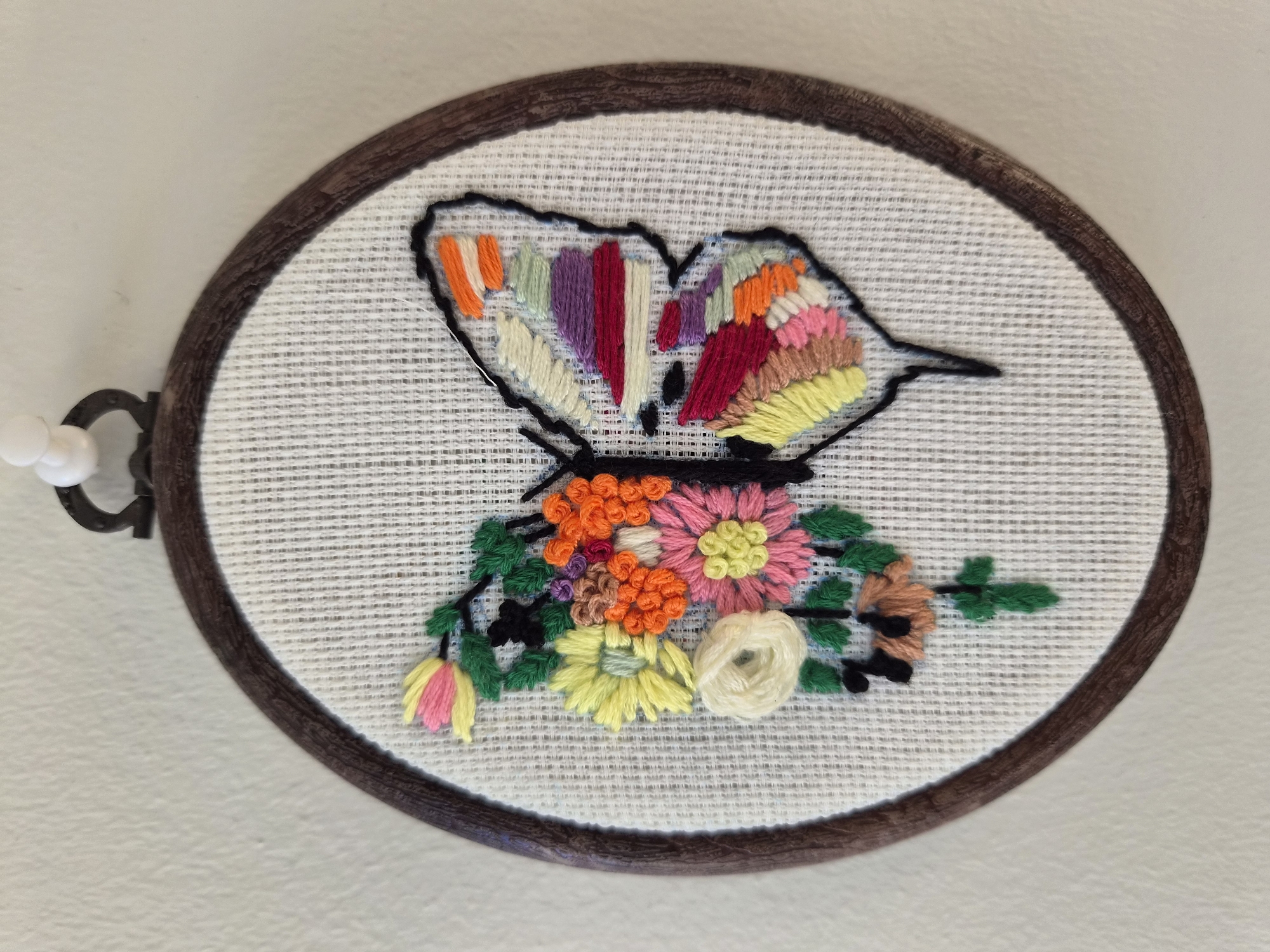 An oval embroidery hoop hangs on a white wall, secured by a white pushpin. The hoop contains a circular piece of white Aida cloth, upon which a butterfly design is embroidered. The butterfly's left half is depicted with embroidered flowers and leaves in various colors, including yellow, pink, orange, green, and brown. The right half of the butterfly's wings features colorful striped embroidery in shades of orange, white, purple, red, light green, and yellow. The butterfly's body and outline are stitched in black thread. The scene is lit from the front, casting minimal shadows. There are no people or animals present in the image. The location is described as being in the city of Şile, Turkey, though this information is not visually discernible from the image itself.