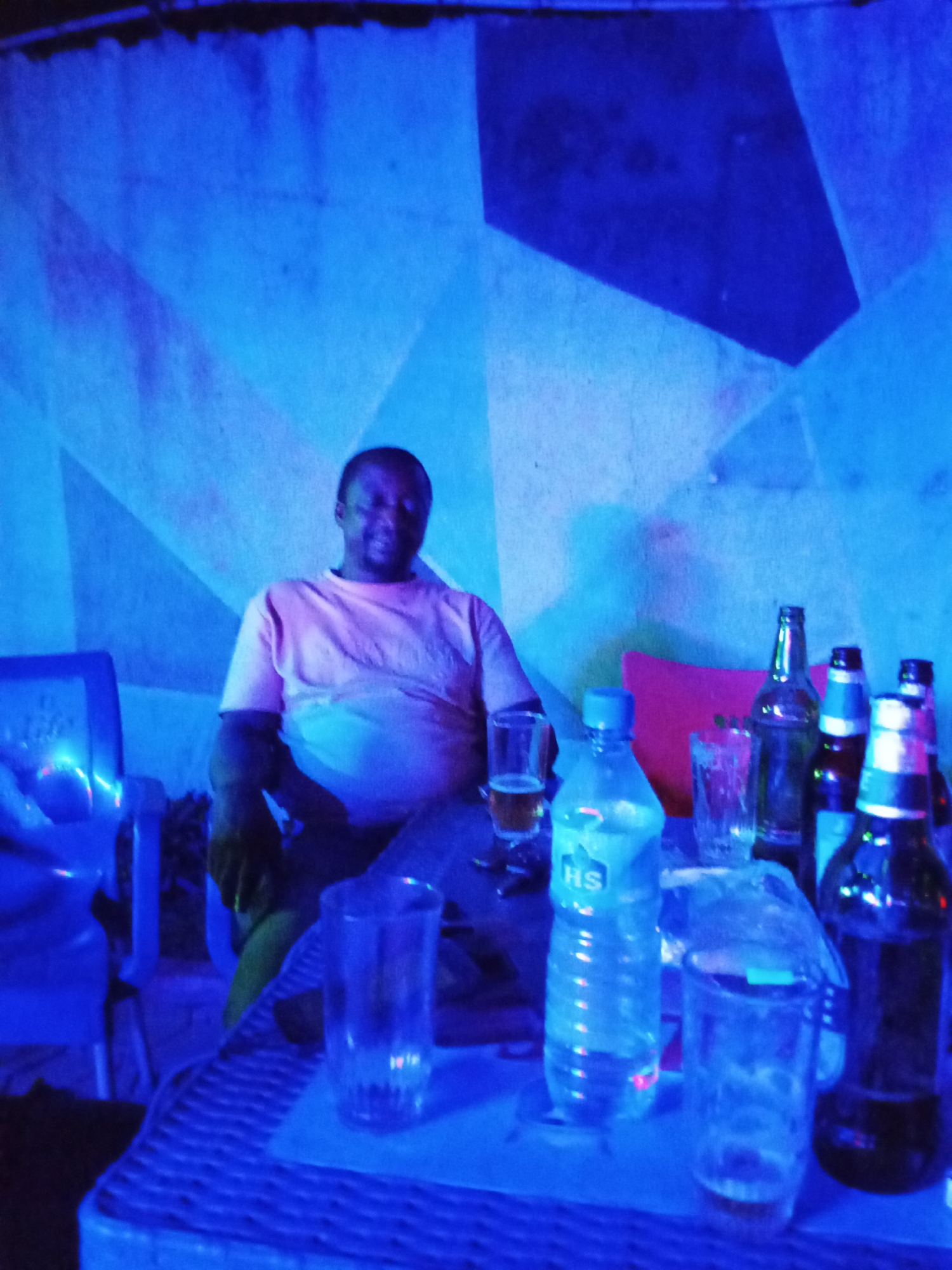 A man in a pink shirt sits at a table in a dimly lit room, bathed in blue light. He is seated to the left of the frame, facing forward with a slight smile. On the table in front of him are several bottles and glasses. From left to right, there is a glass with a golden liquid, a plastic water bottle, an empty glass, and several beer bottles. The background features a wall with an abstract geometric pattern in shades of blue and white. The lighting creates a somber and somewhat surreal atmosphere.