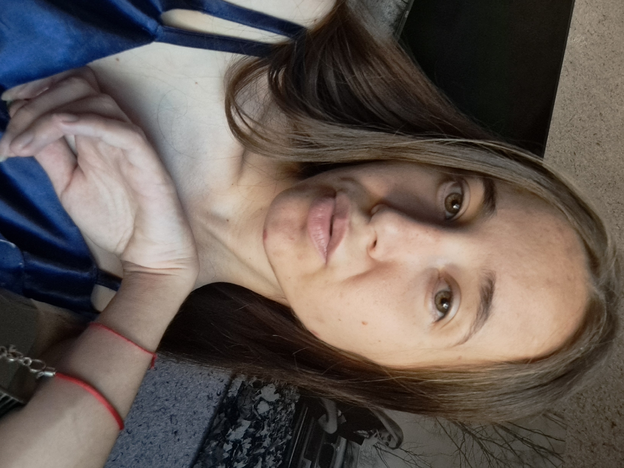 In Vypasne, Ukraine, a young woman with long, brown hair and light skin is captured in a selfie. She is wearing a dark blue top with thin straps. Her right arm is bent, with her hand resting on her chest. A thin red string bracelet is visible on her left wrist, and another red string bracelet is on her right wrist, featuring a silver clasp. The background is blurred, showing a textured wall and a framed picture of a woman with blonde hair sitting near a car. The lighting suggests an indoor setting.